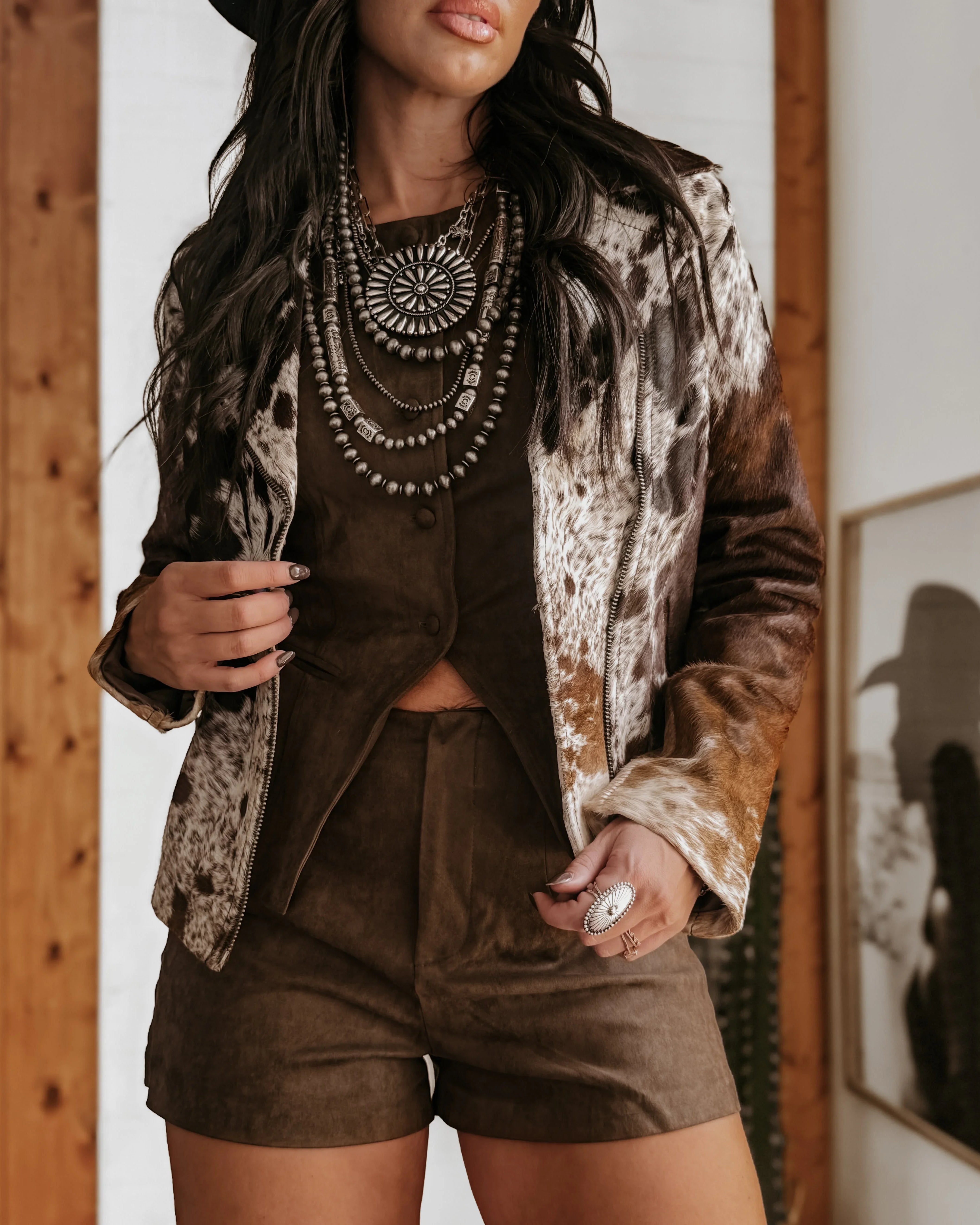 Woman in a western cow print jacket, brown suede shorts, and layered boho necklaces.