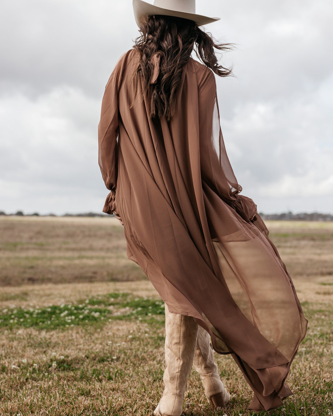 Woman in sheer brown boho duster, cowboy hat and boots in a field, western outfit style