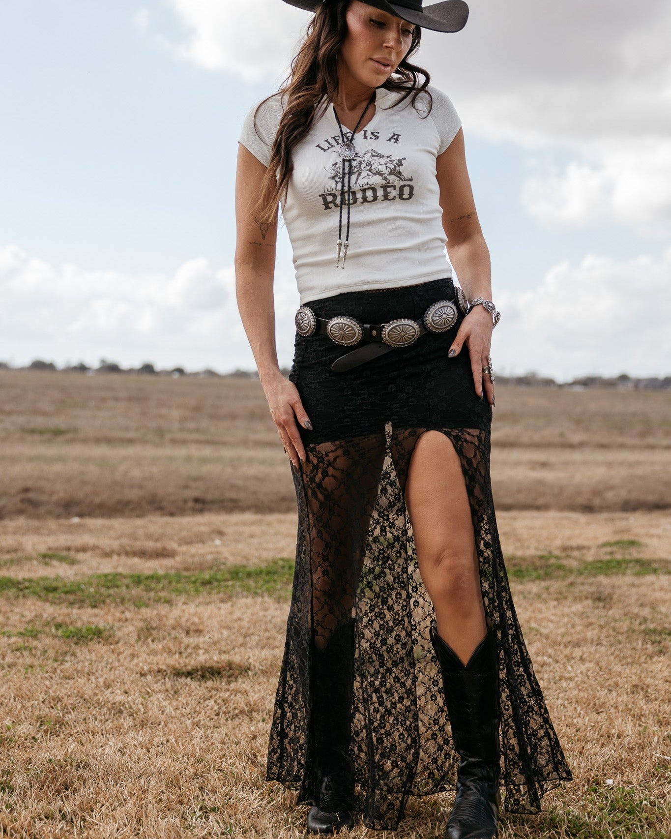 Woman in cowgirl hat, graphic tee, black lace skirt with slit, concho belt and western boots outdoors