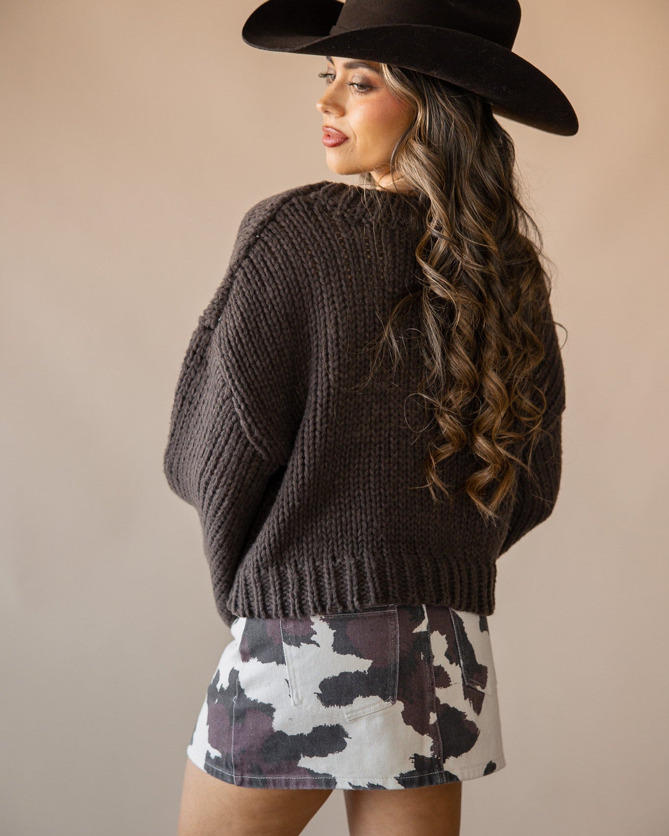 Woman in a western hat, chunky knit sweater, cow print skirt, and suede boots