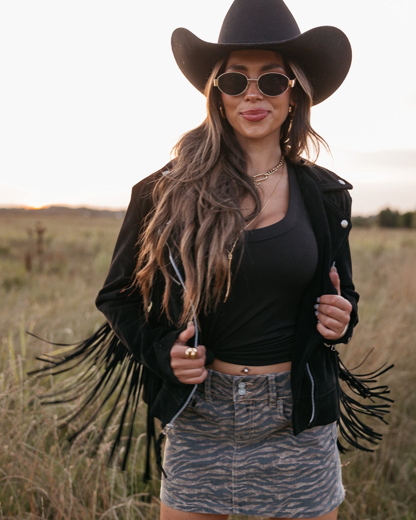 Woman wearing a black cowboy hat, fringe jacket, and sunglasses in a field.