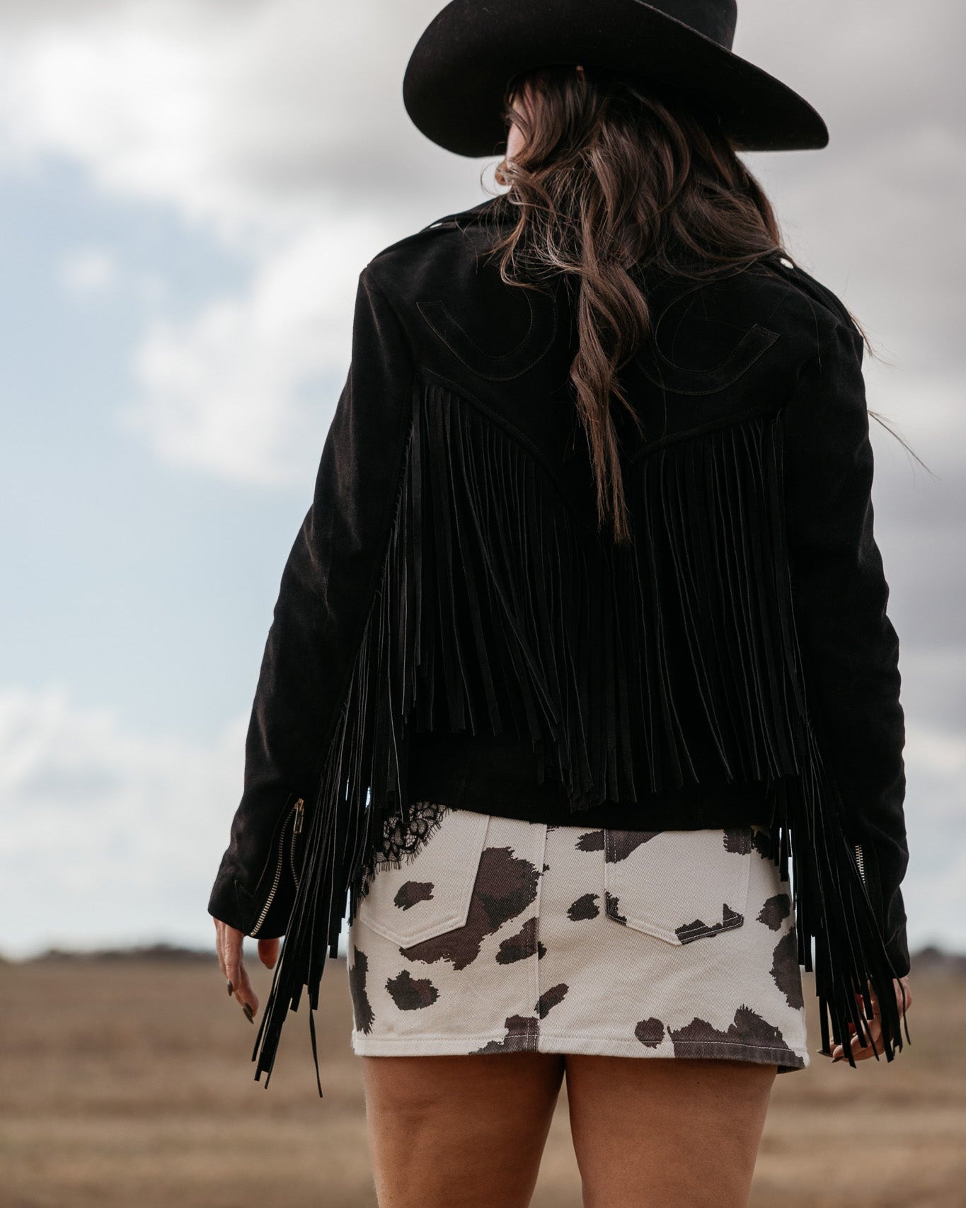 Woman in black fringe jacket and cow print skirt wearing cowboy hat in western landscape
