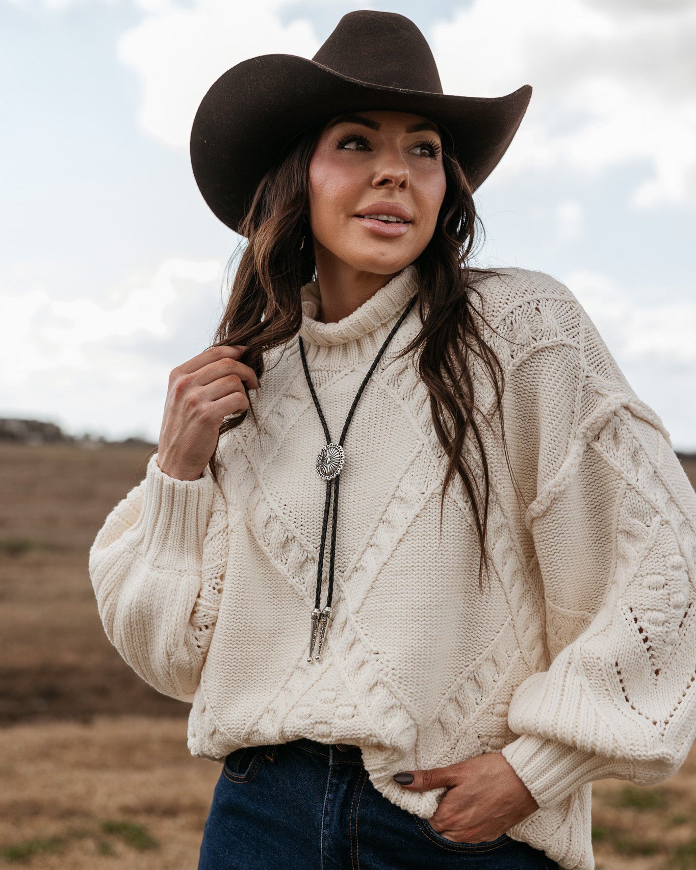 Woman in a black cowboy hat, cream knit sweater, denim jeans, and bolo tie outdoors
