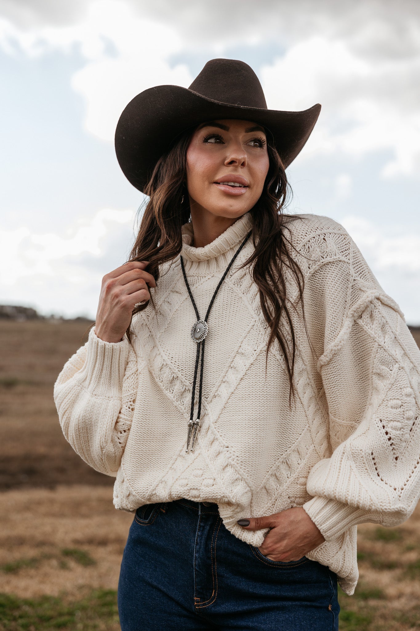 Woman in a black cowboy hat, cream knit sweater, denim jeans, and bolo tie outdoors