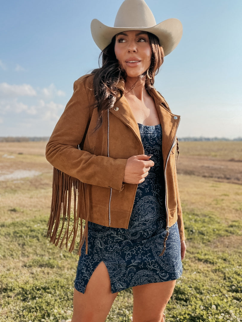 Woman in a suede fringe jacket, paisley dress, and cowboy hat in a western outdoor setting