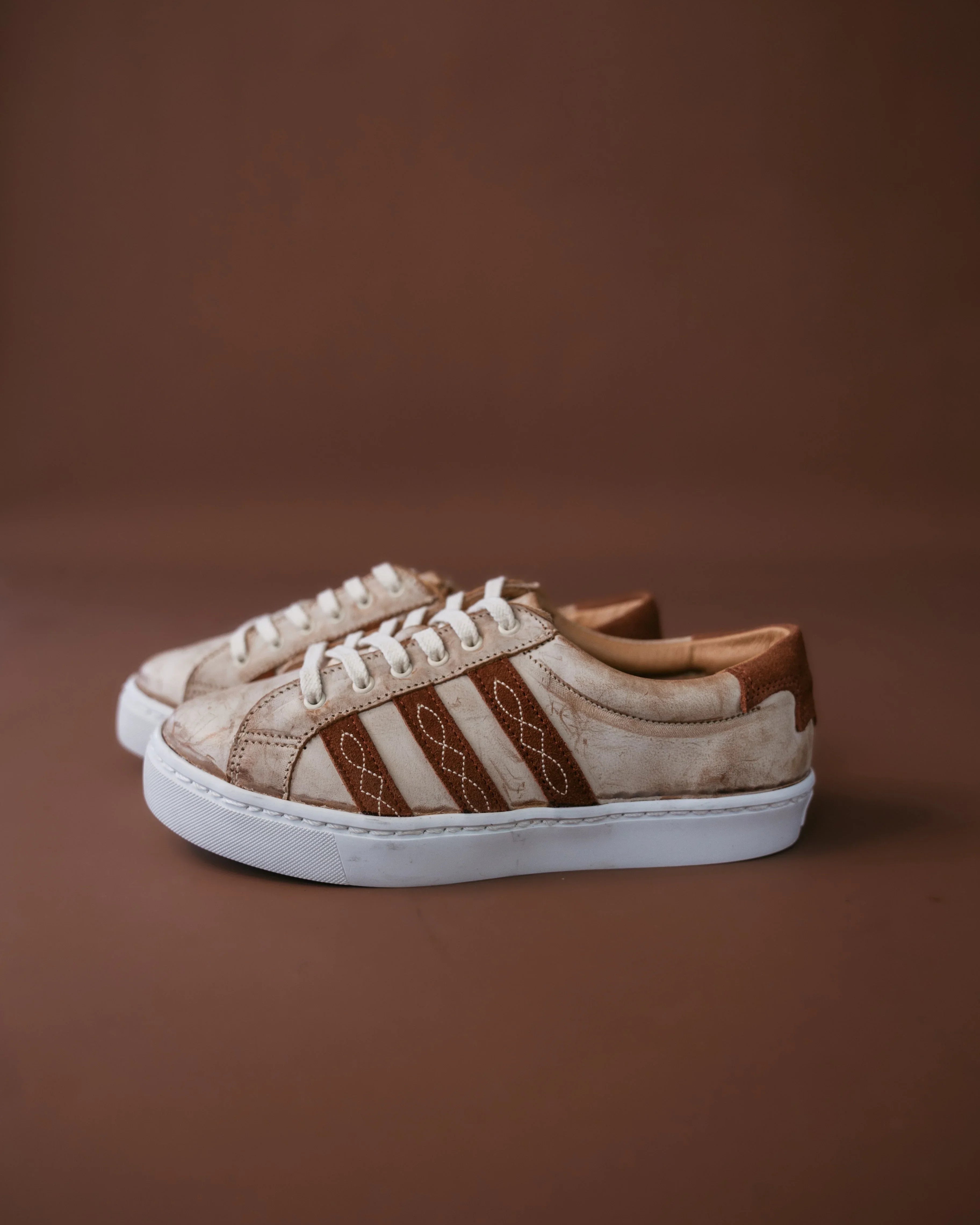 Beige and brown western sneakers with stitched detail, white sole, on brown background