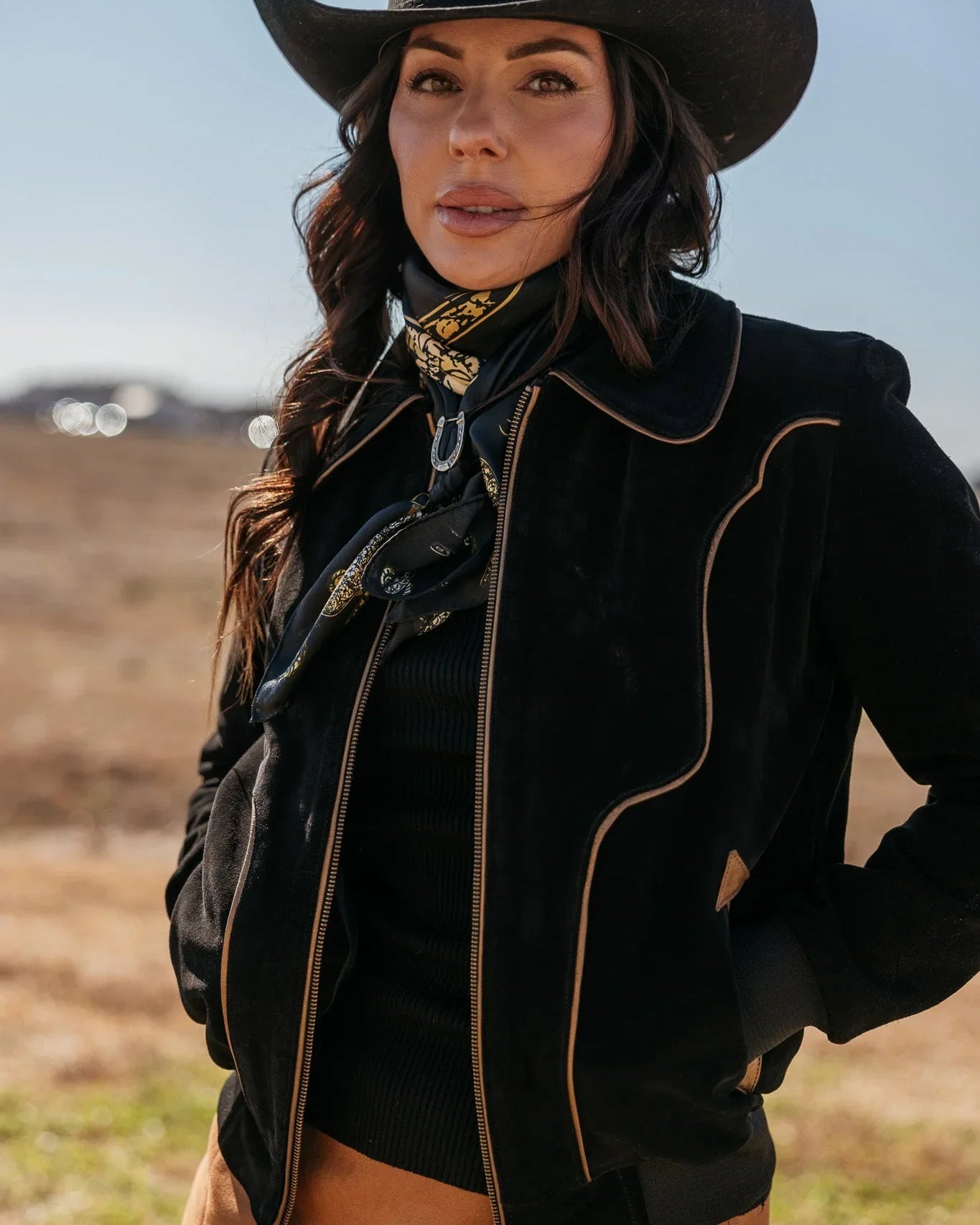 Woman in western boho outfit with black cowgirl hat, suede jacket, and bandana outdoors