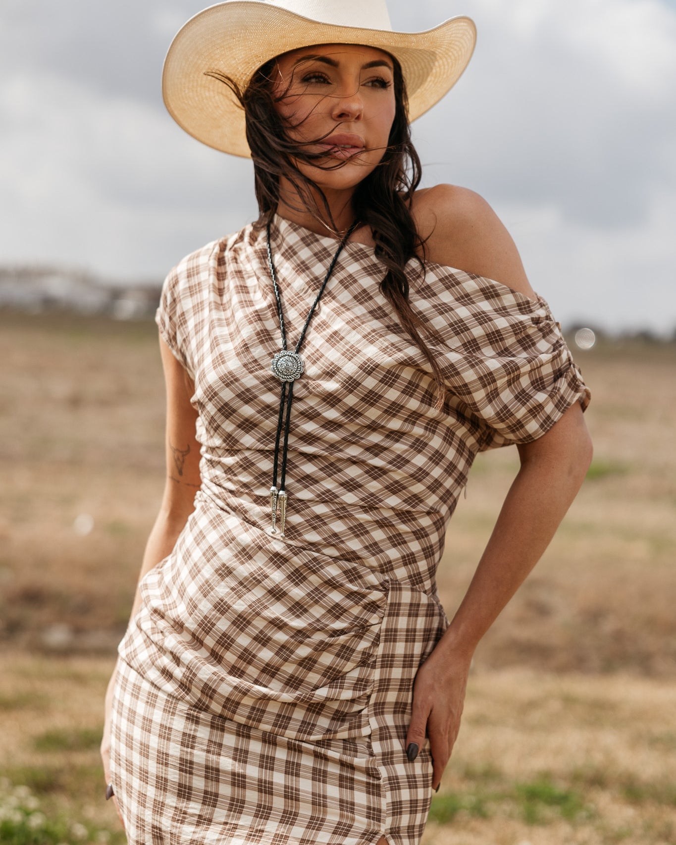 Woman in a brown plaid western dress, cowboy hat, and bolo tie standing outdoors