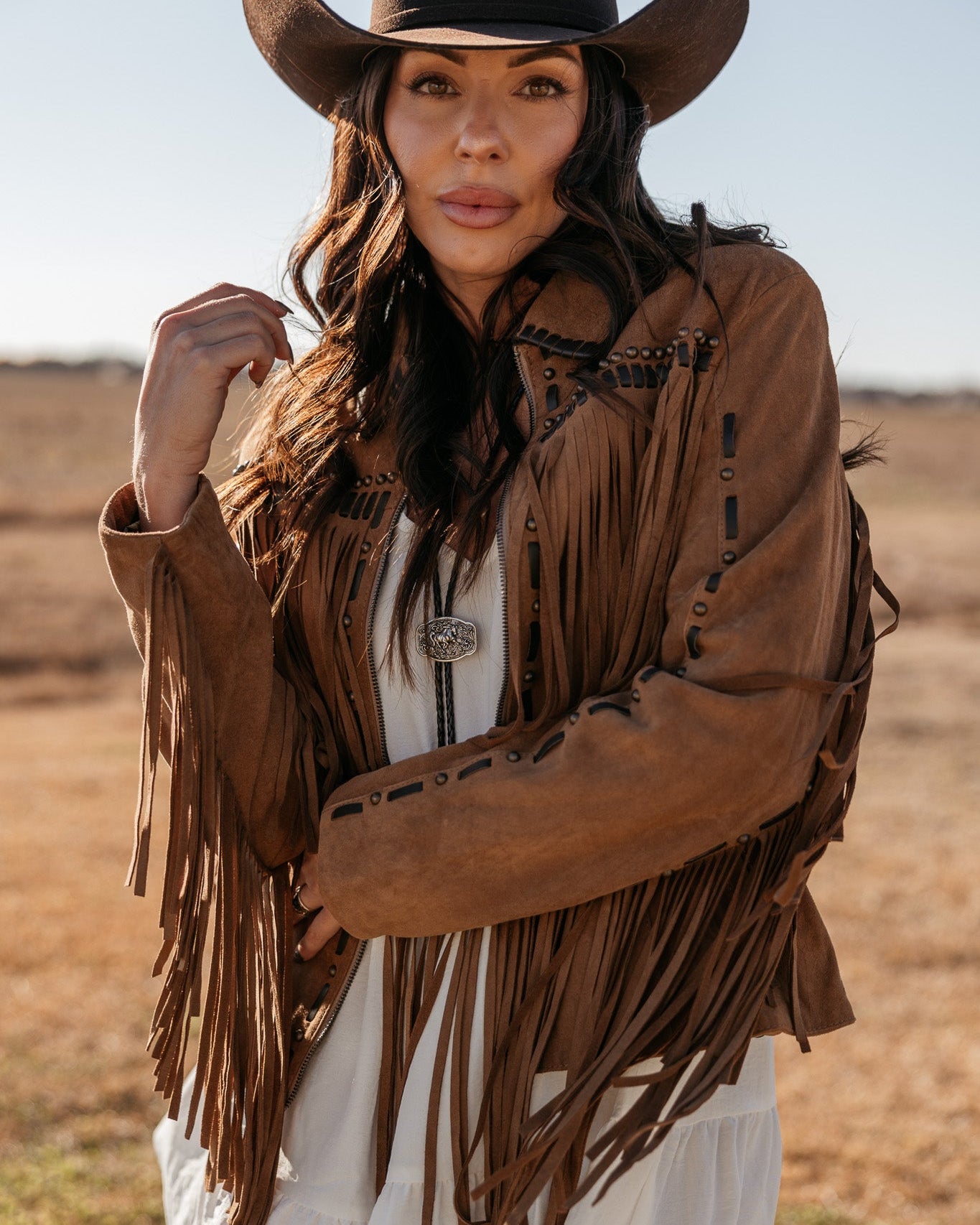 Woman in a brown fringe suede jacket and black cowboy hat in a western outdoor setting