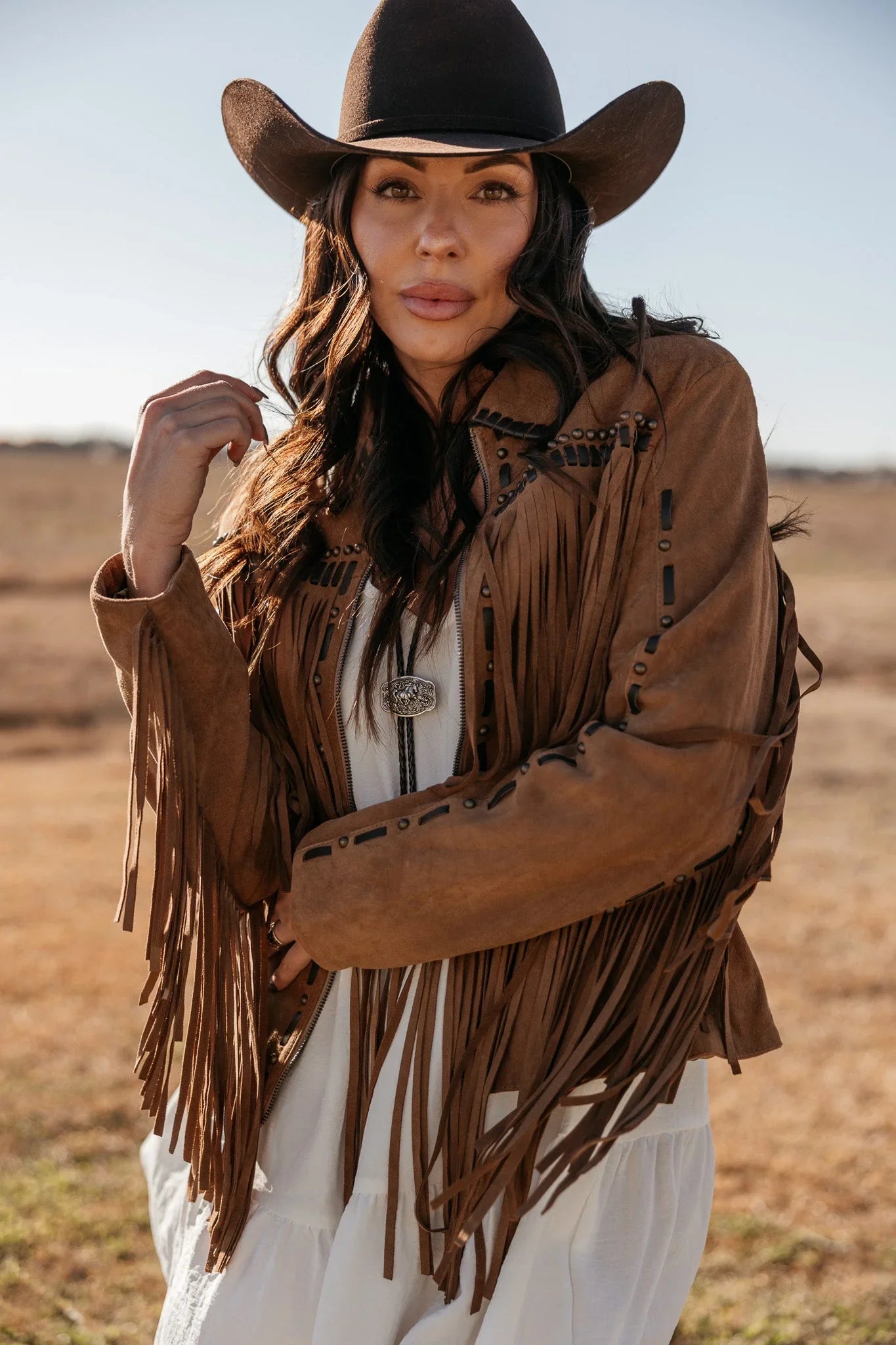 Woman in a brown fringe suede jacket and black cowboy hat in a western outdoor setting