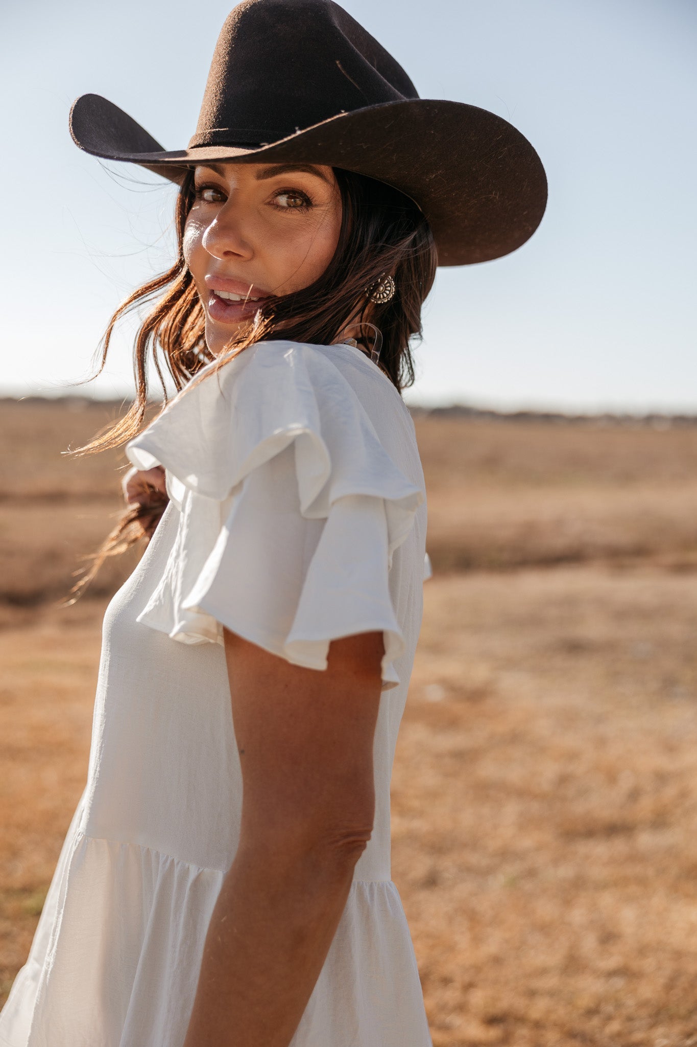 Woman in white ruffle dress and brown cowboy hat in a sunny western field