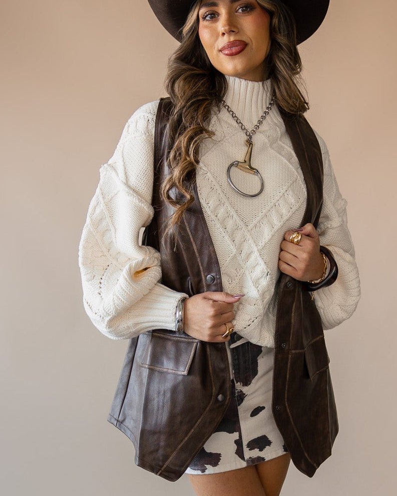 Woman in cowgirl hat, leather vest, cream sweater, and cow print skirt styled western boho