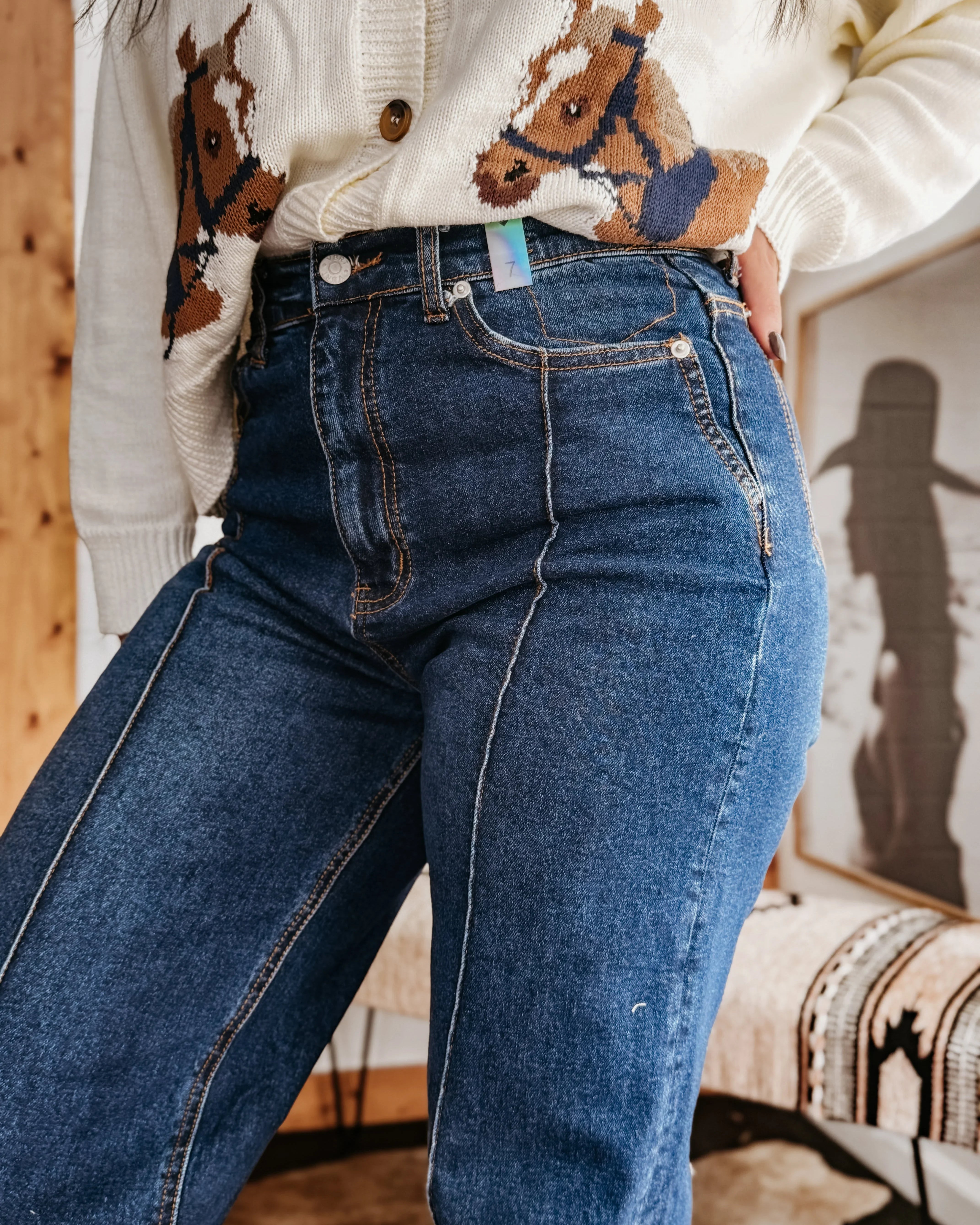 Woman in high-waisted western jeans and cream sweater with horse print, boho decor background