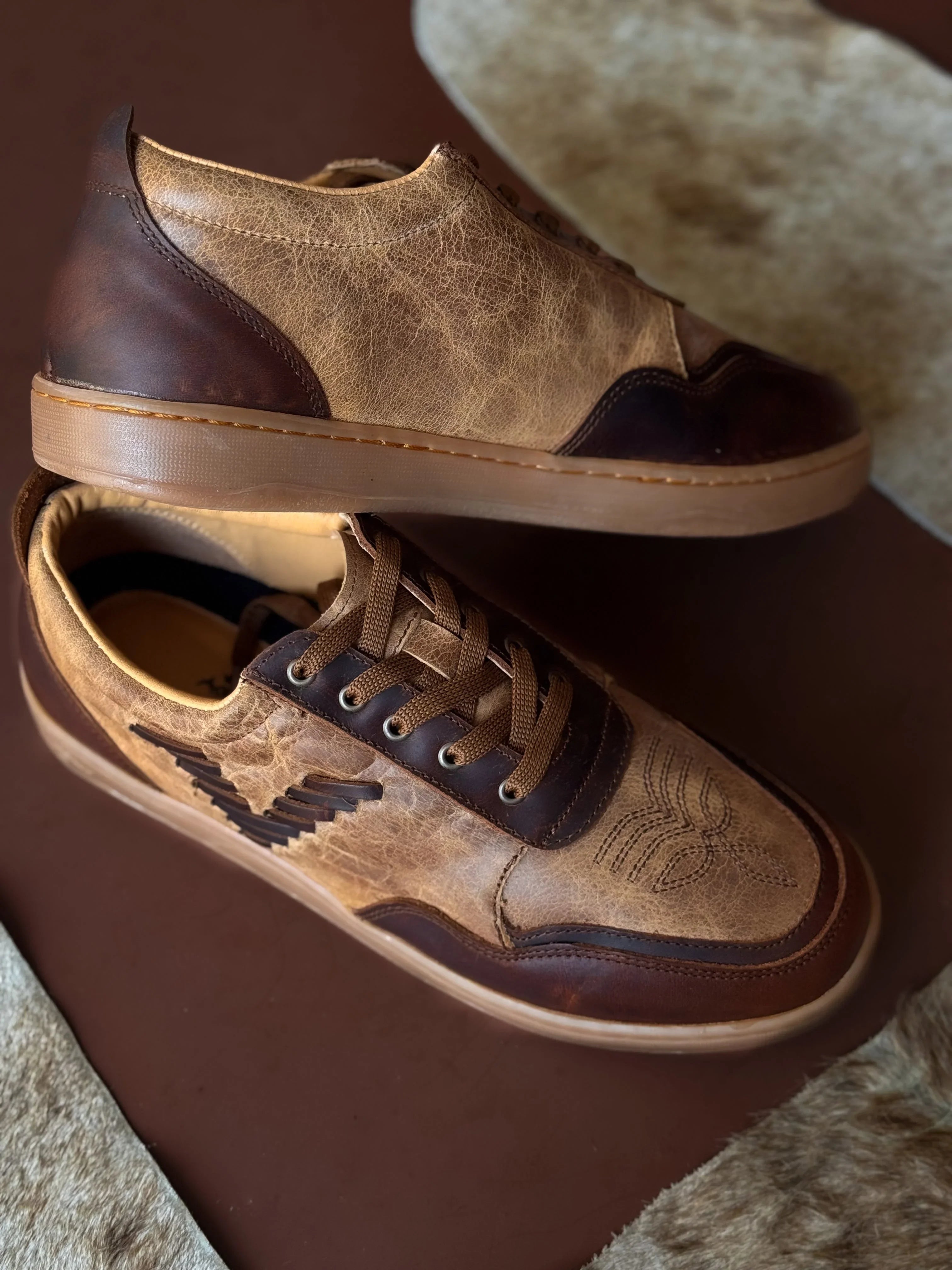 Western-style brown leather sneakers with stitched detailing on a cowhide and brown background