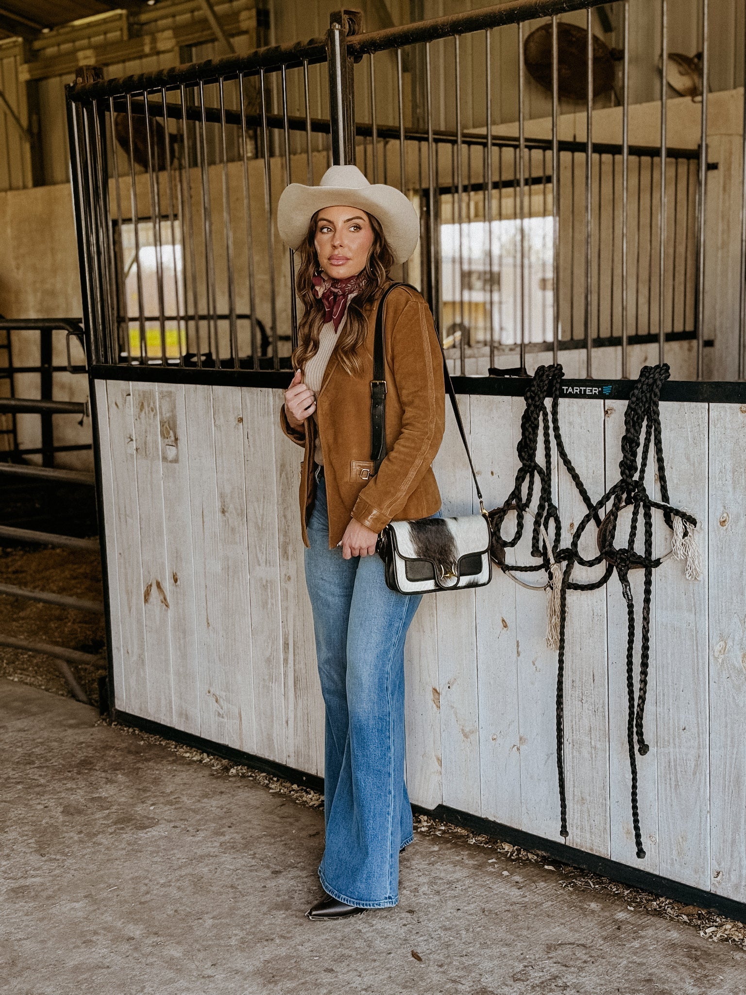 Woman in western boho outfit with suede jacket, cowboy hat, flared jeans in a horse stable