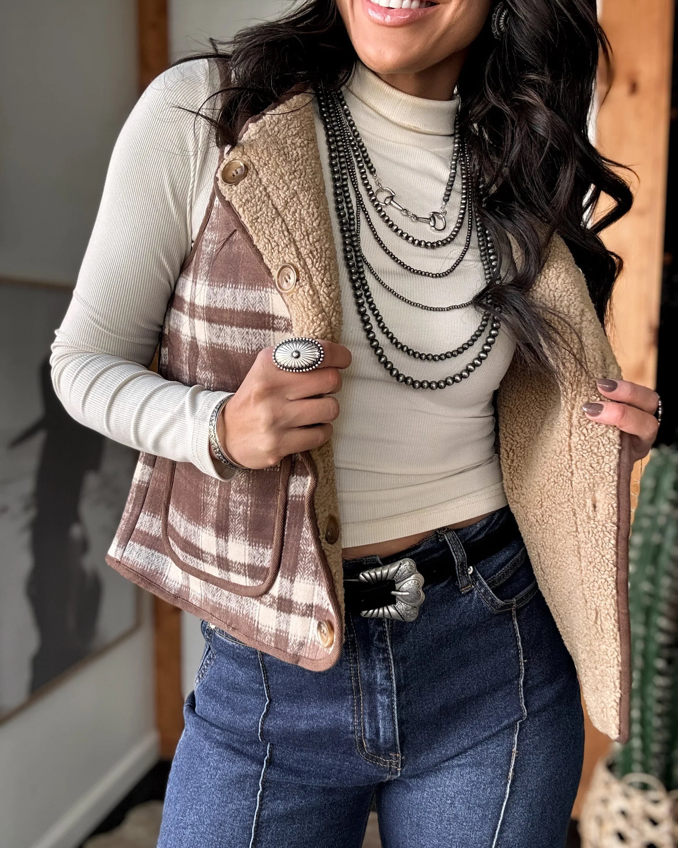 Woman wearing a plaid vest over a turtleneck with layered necklaces, smiling indoors.