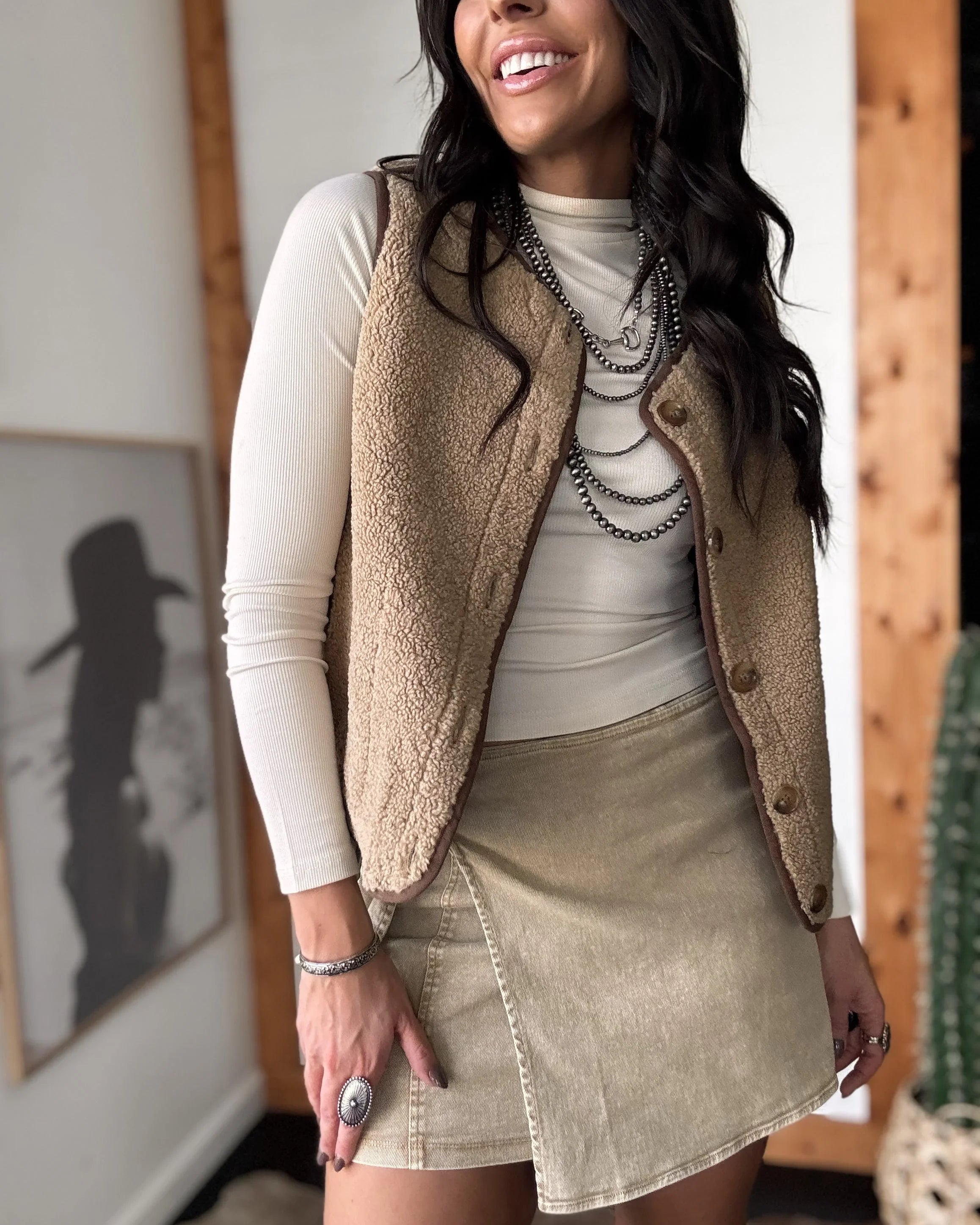 Woman wearing a beige vest over a white long-sleeve shirt and tan skirt indoors.