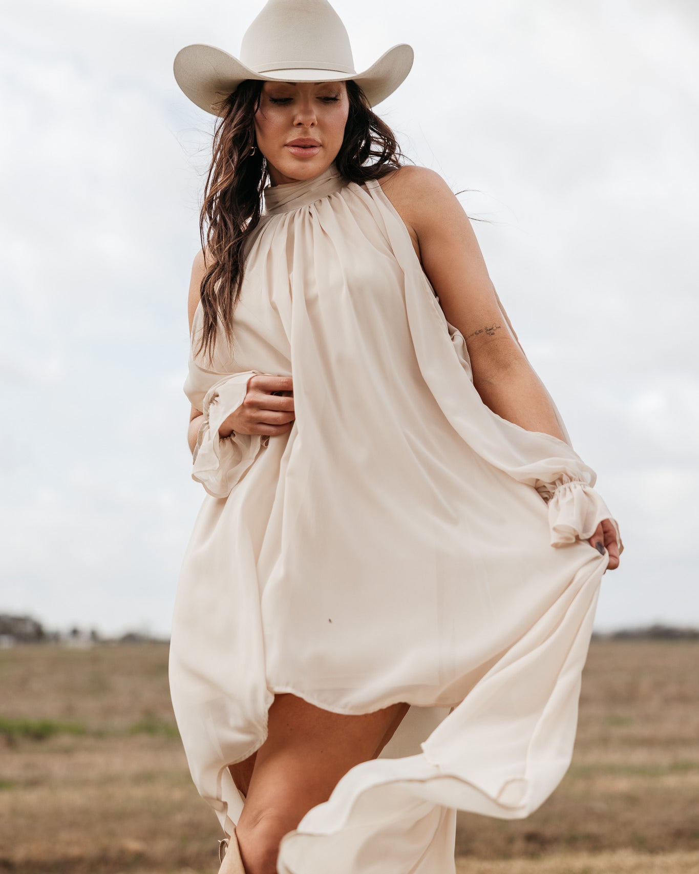 Woman in cream western dress and cowboy hat posing outdoors in boho cowgirl style