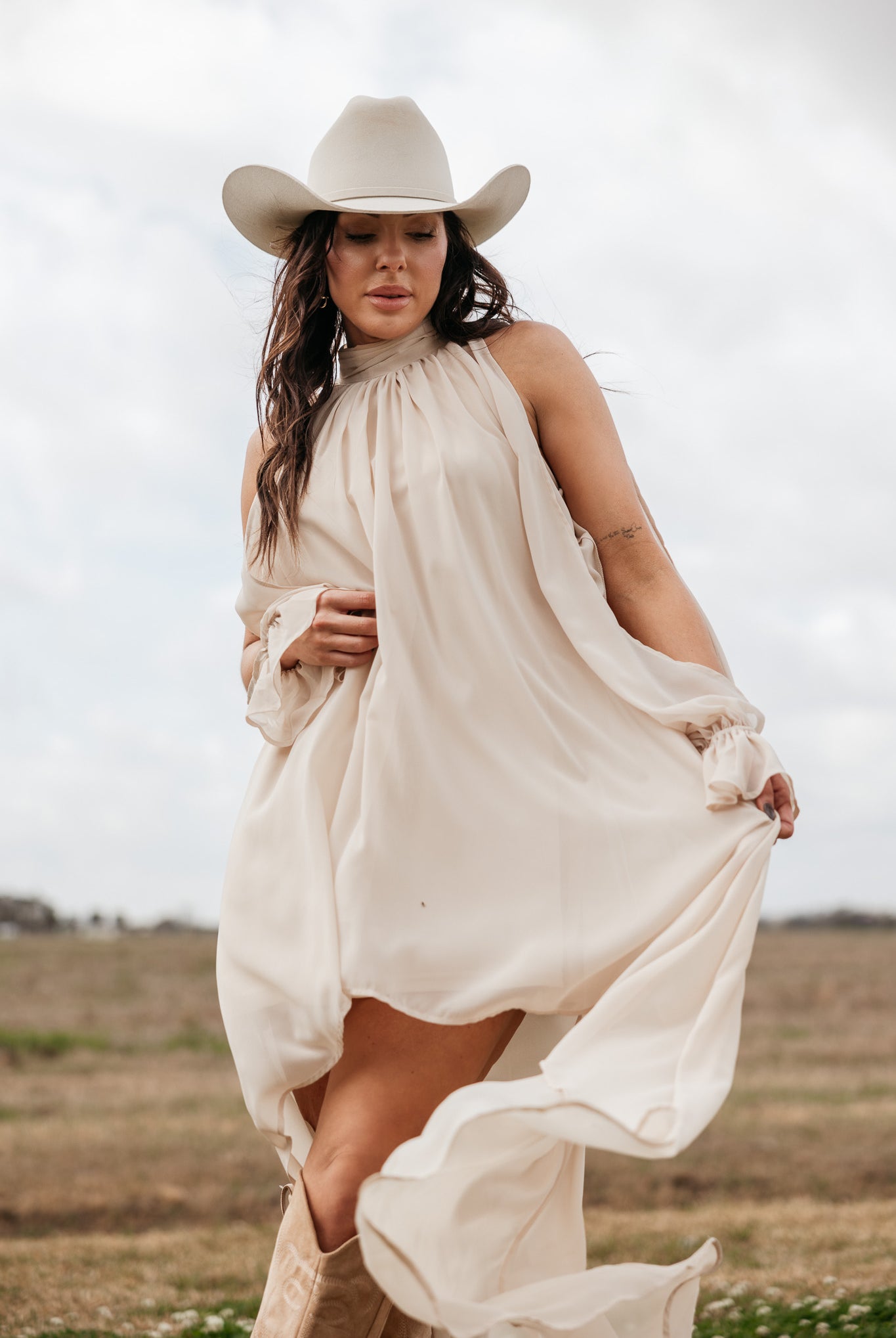 Woman in cream western dress and cowboy hat posing outdoors in boho cowgirl style