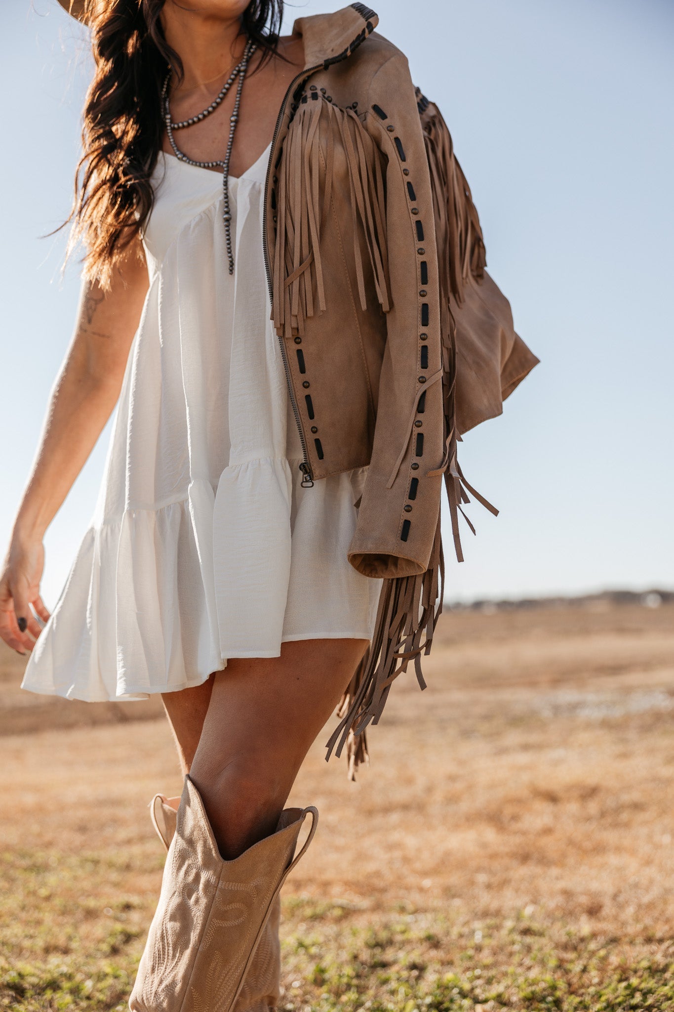 Woman in white boho dress, fringe suede jacket, and western boots outdoors