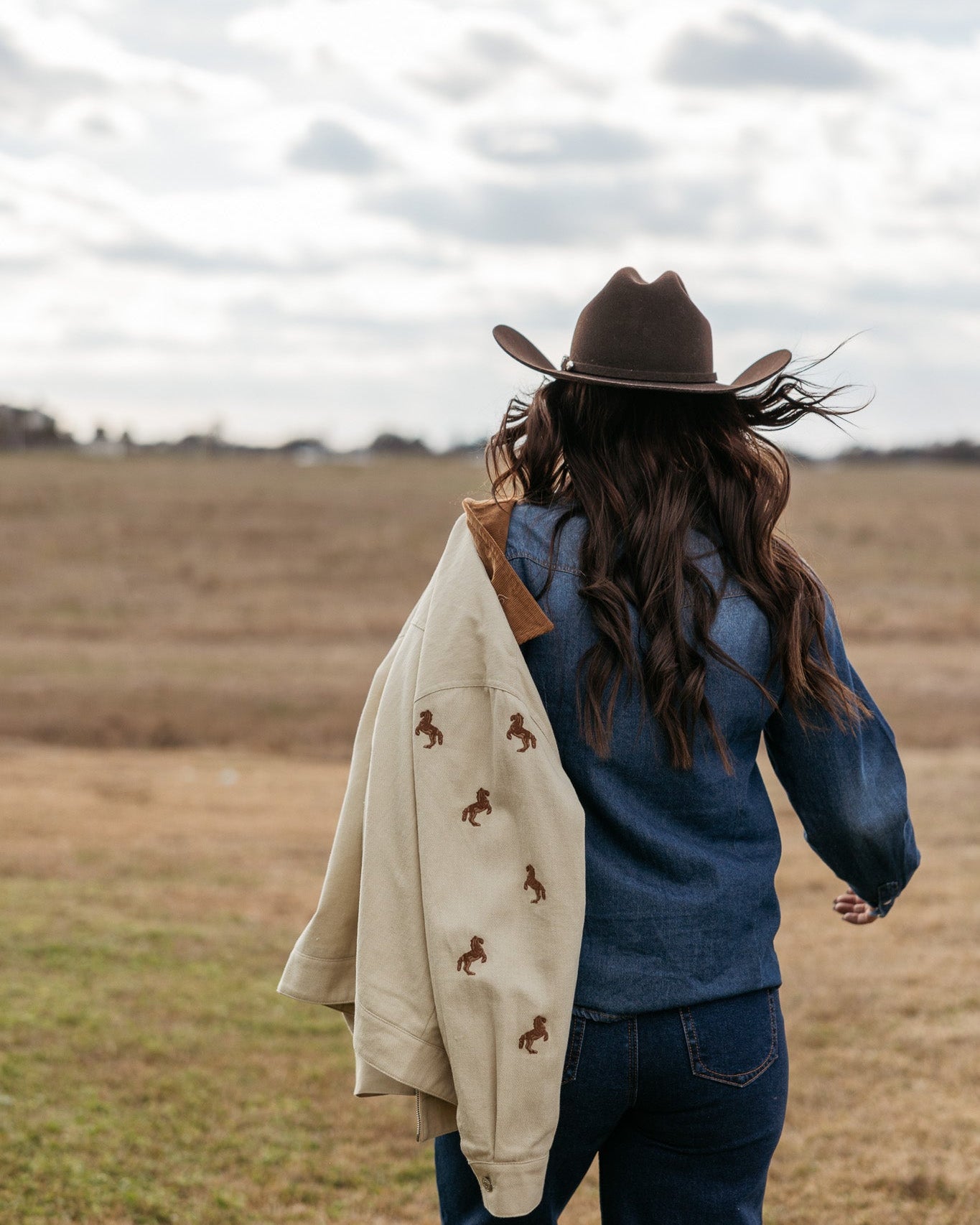 Woman in cowboy hat and denim outfit outdoors, holding tan jacket with horse embroidery, western style