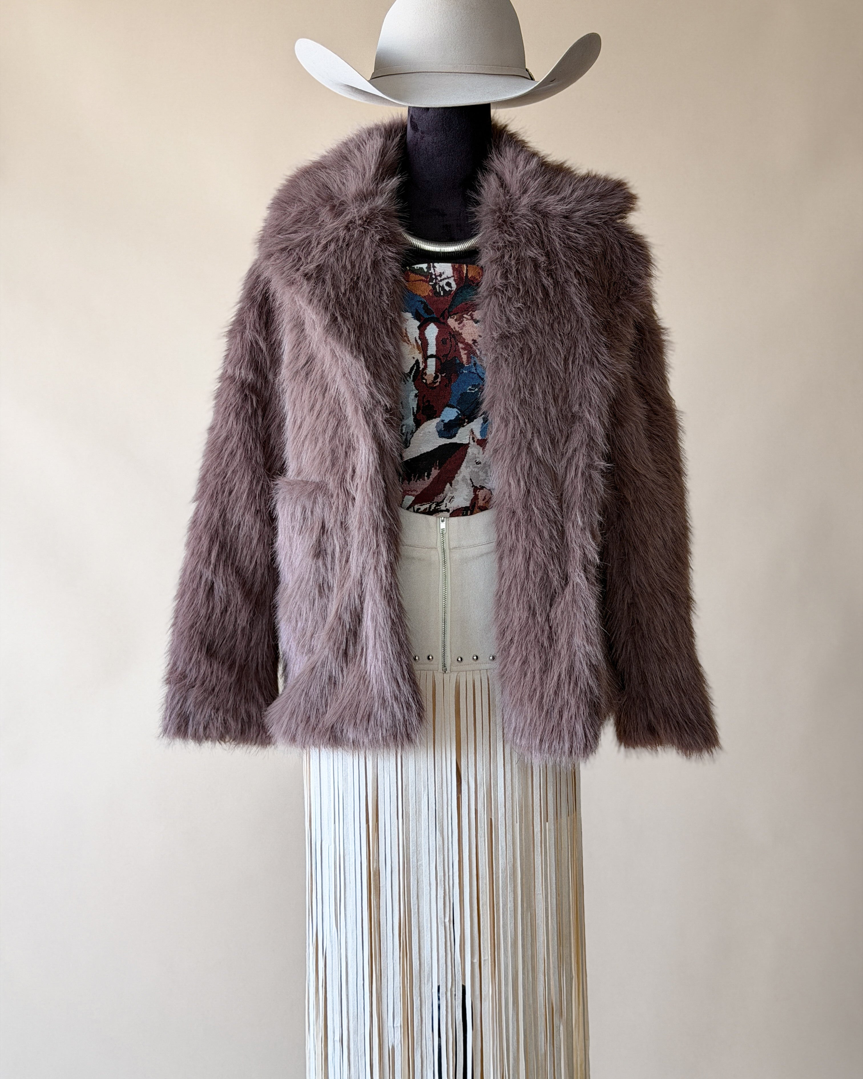 Western cowgirl outfit with mauve faux fur jacket, graphic tee, white fringe skirt, and wide-brim hat