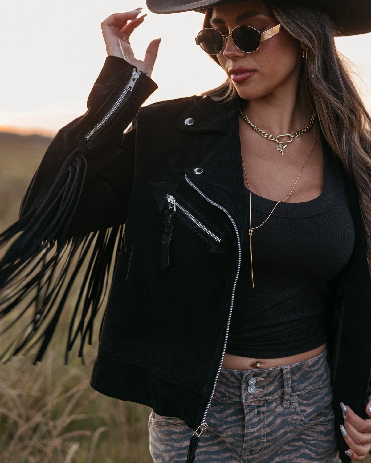 Woman in a black fringe jacket, black top, and denim skirt with a cowboy hat in a field.