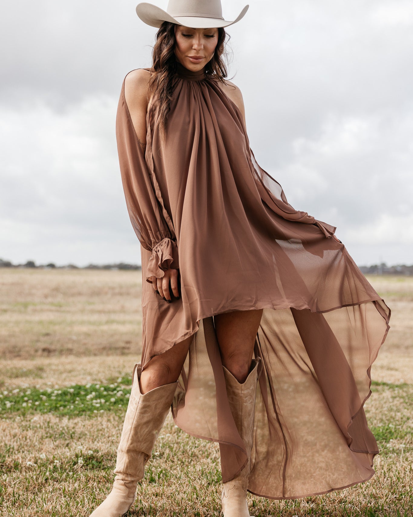 Woman in flowy brown western dress, beige cowboy hat, and boots standing in a field
