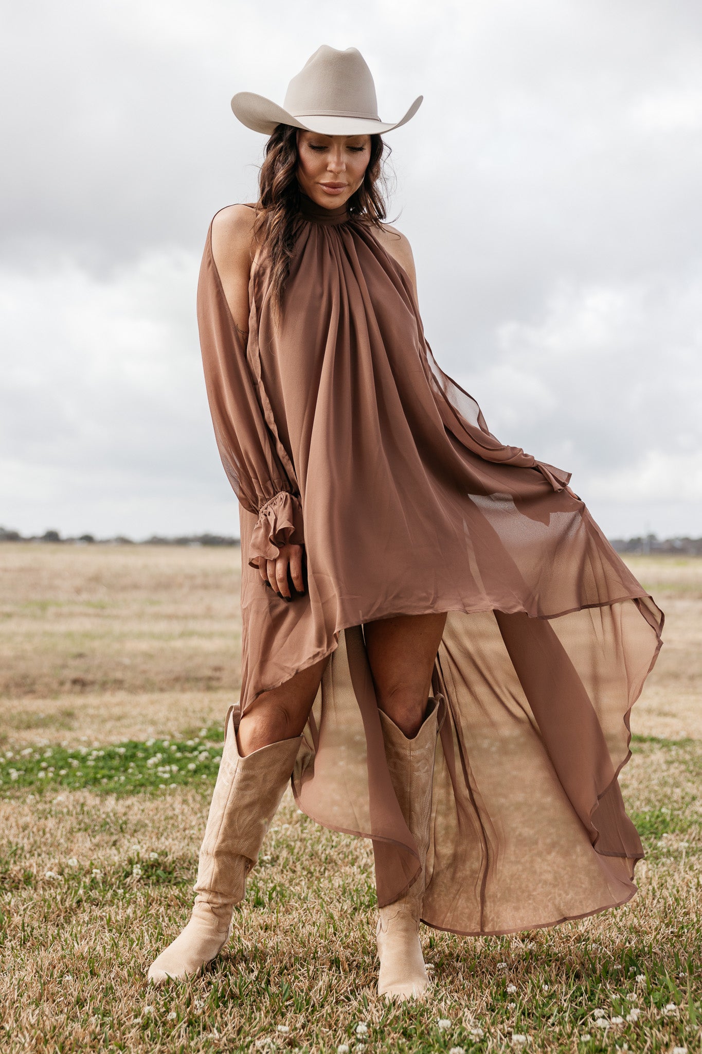 Woman in flowy brown western dress, beige cowboy hat, and boots standing in a field
