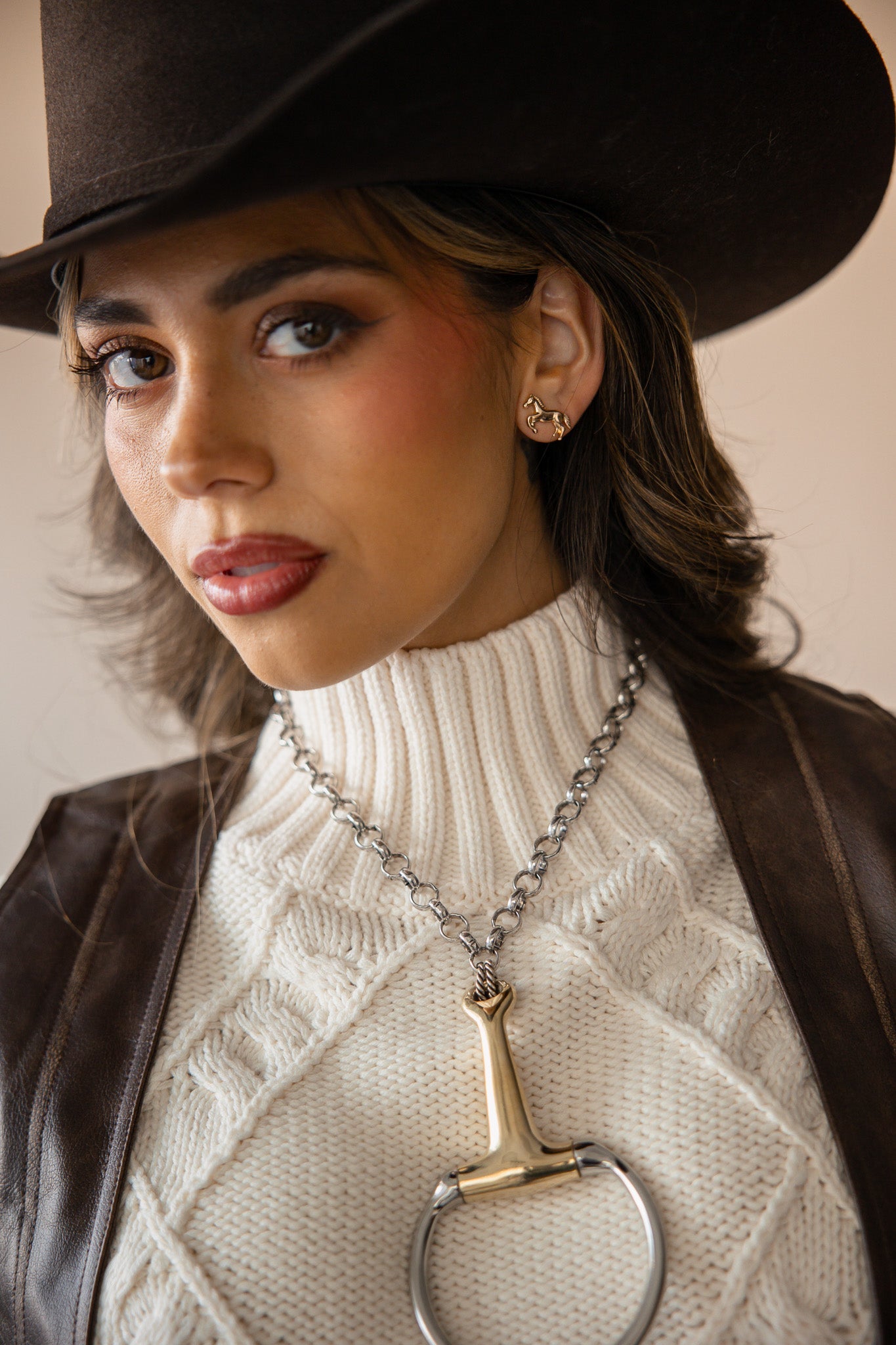 Woman in cowgirl hat, cream sweater, leather vest, with Western jewelry and horse earrings