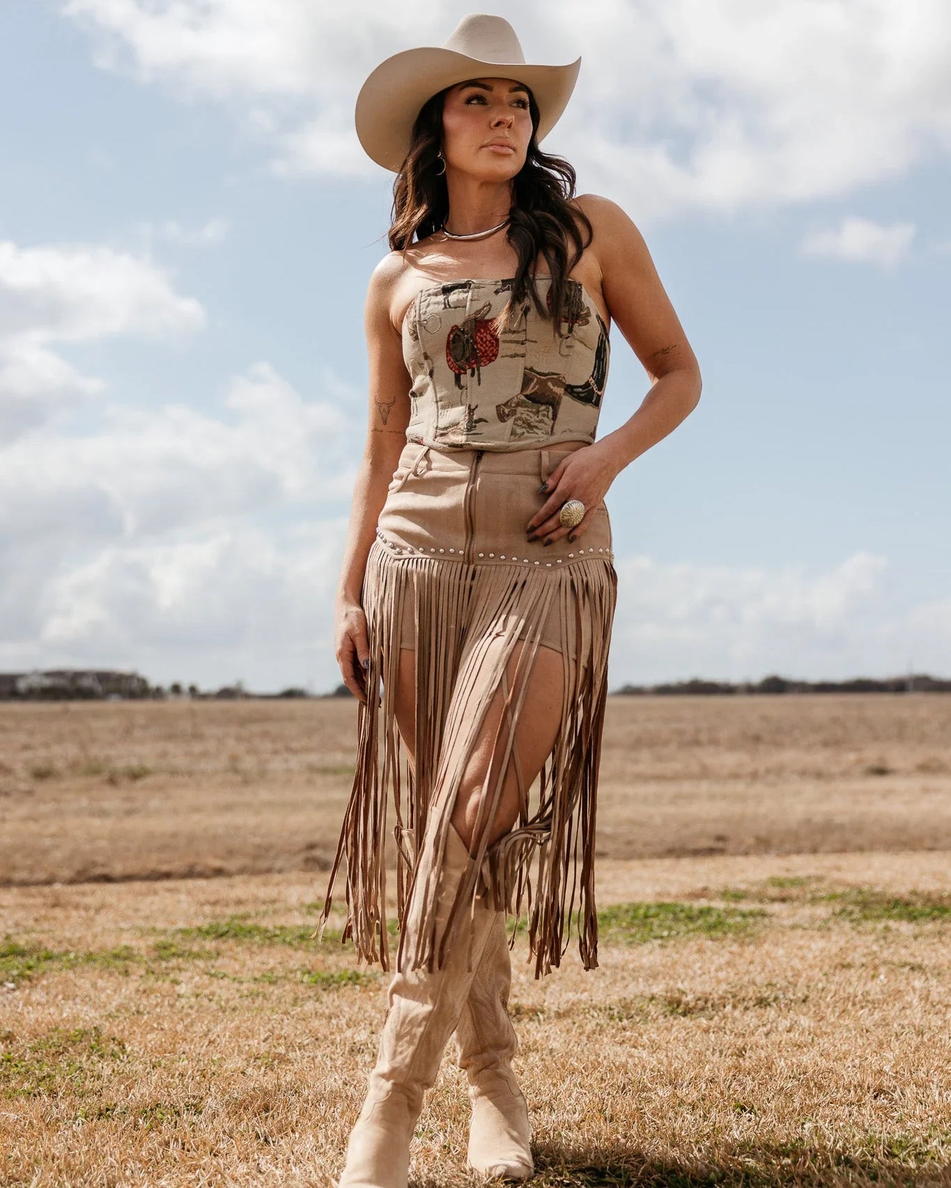 Woman in western boho outfit with beige cowboy hat, fringe skirt, and boots standing outdoors