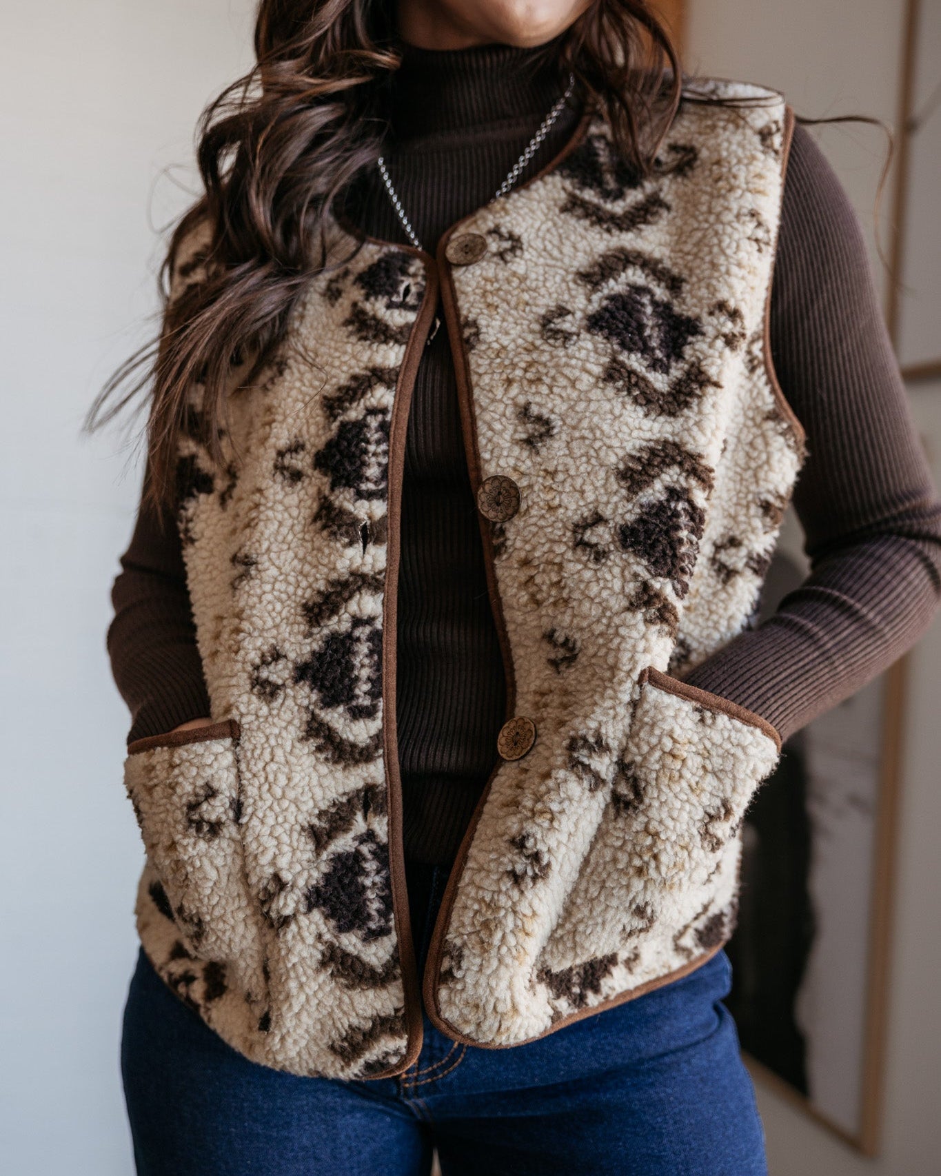 Woman wearing a western sherpa vest with Aztec pattern, brown turtleneck, and blue jeans.