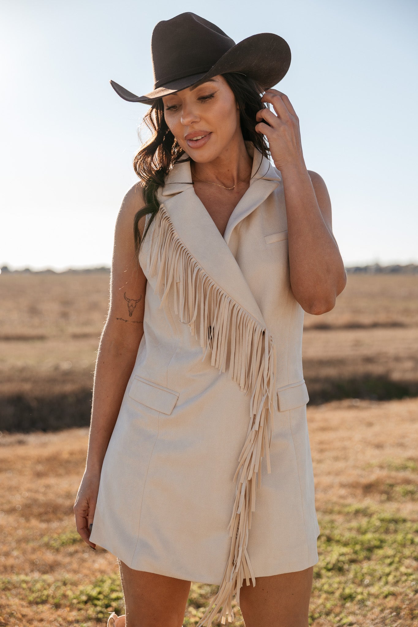 Woman in cream fringe western dress and brown cowgirl hat standing in a field