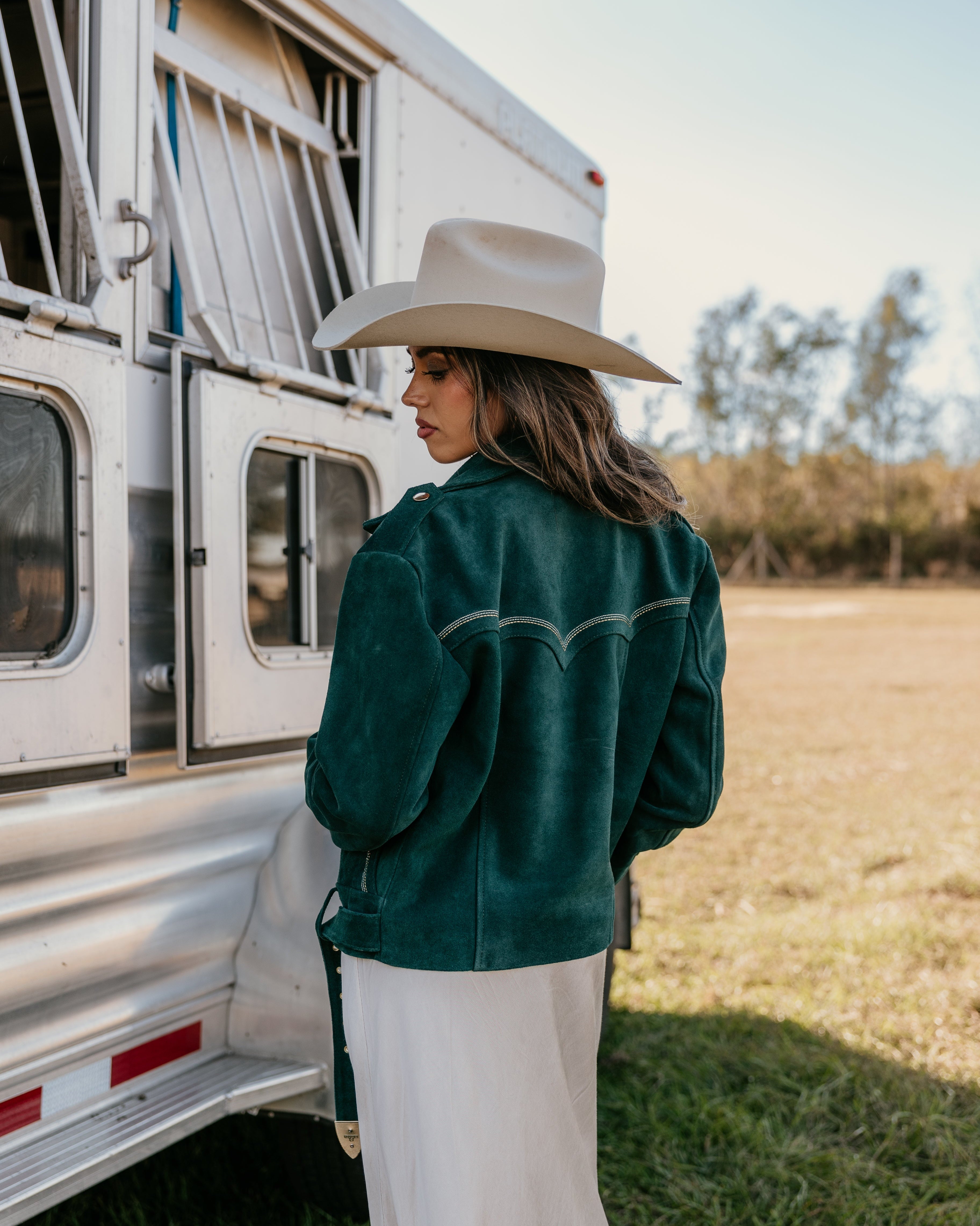 Woman in a wide-brim hat and emerald western jacket by a horse trailer, outdoor boho style