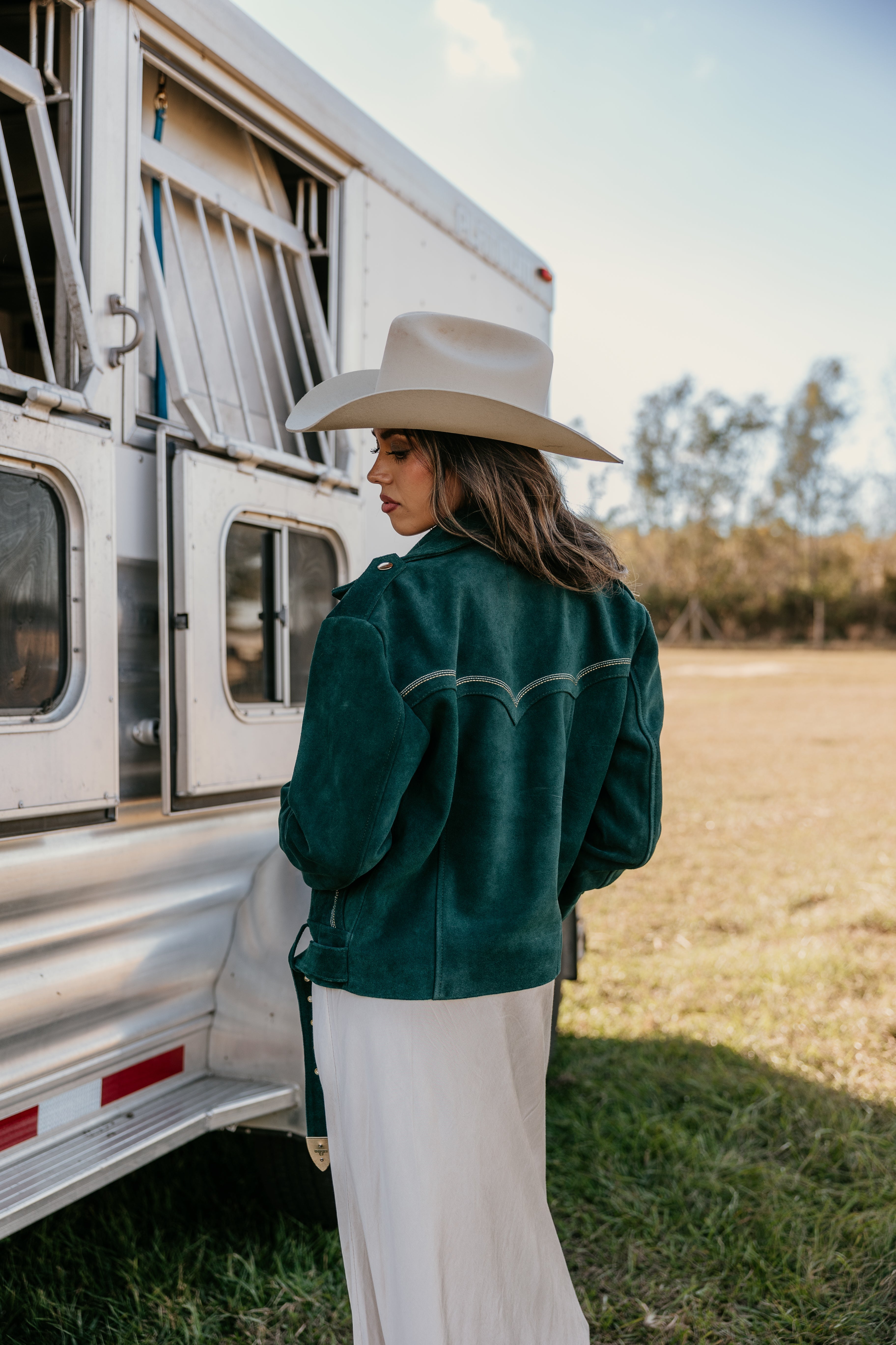 Woman in a wide-brim hat and emerald western jacket by a horse trailer, outdoor boho style
