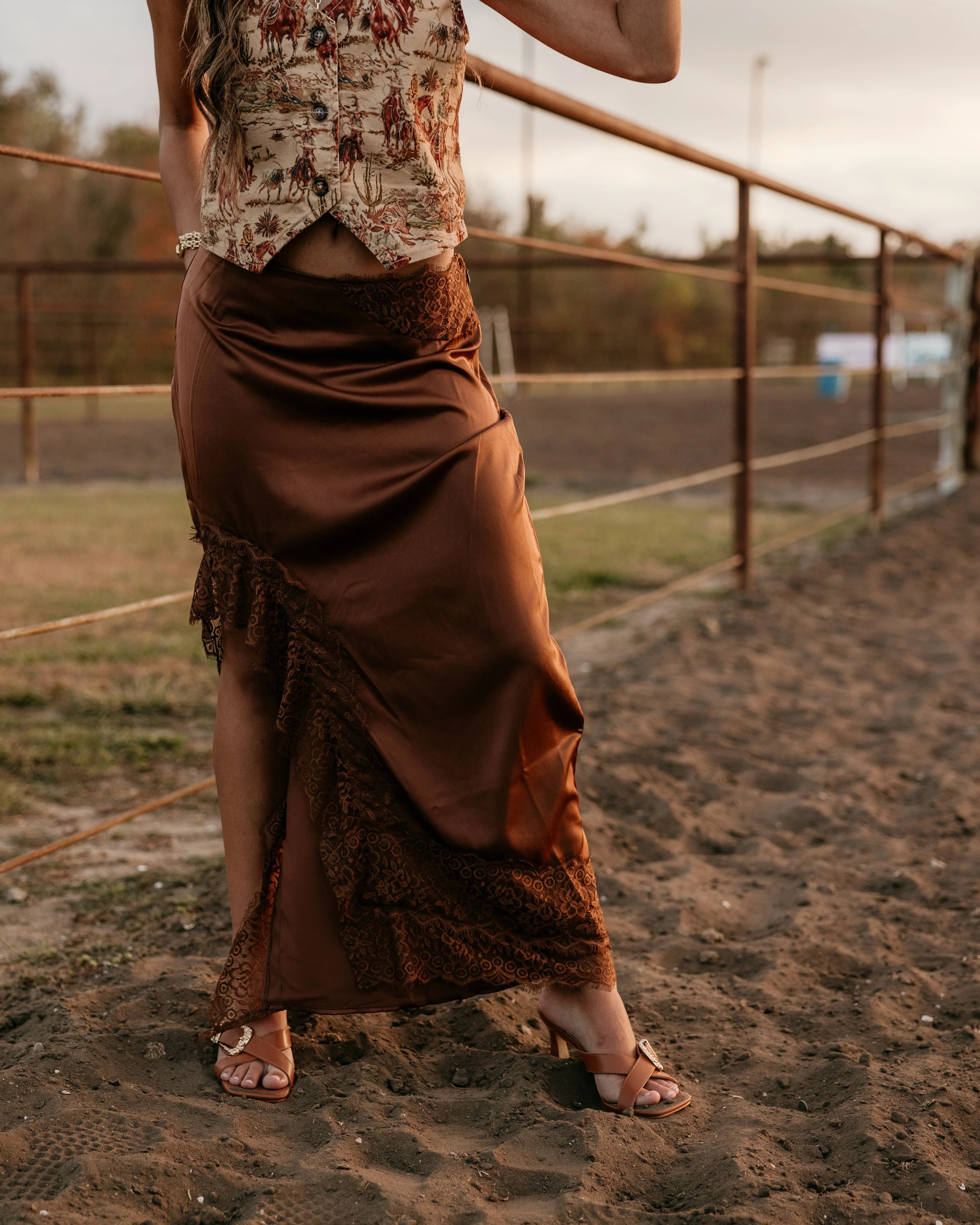 Person wearing a patterned top and brown skirt standing on a dirt path with a fence and grassy area in the background.