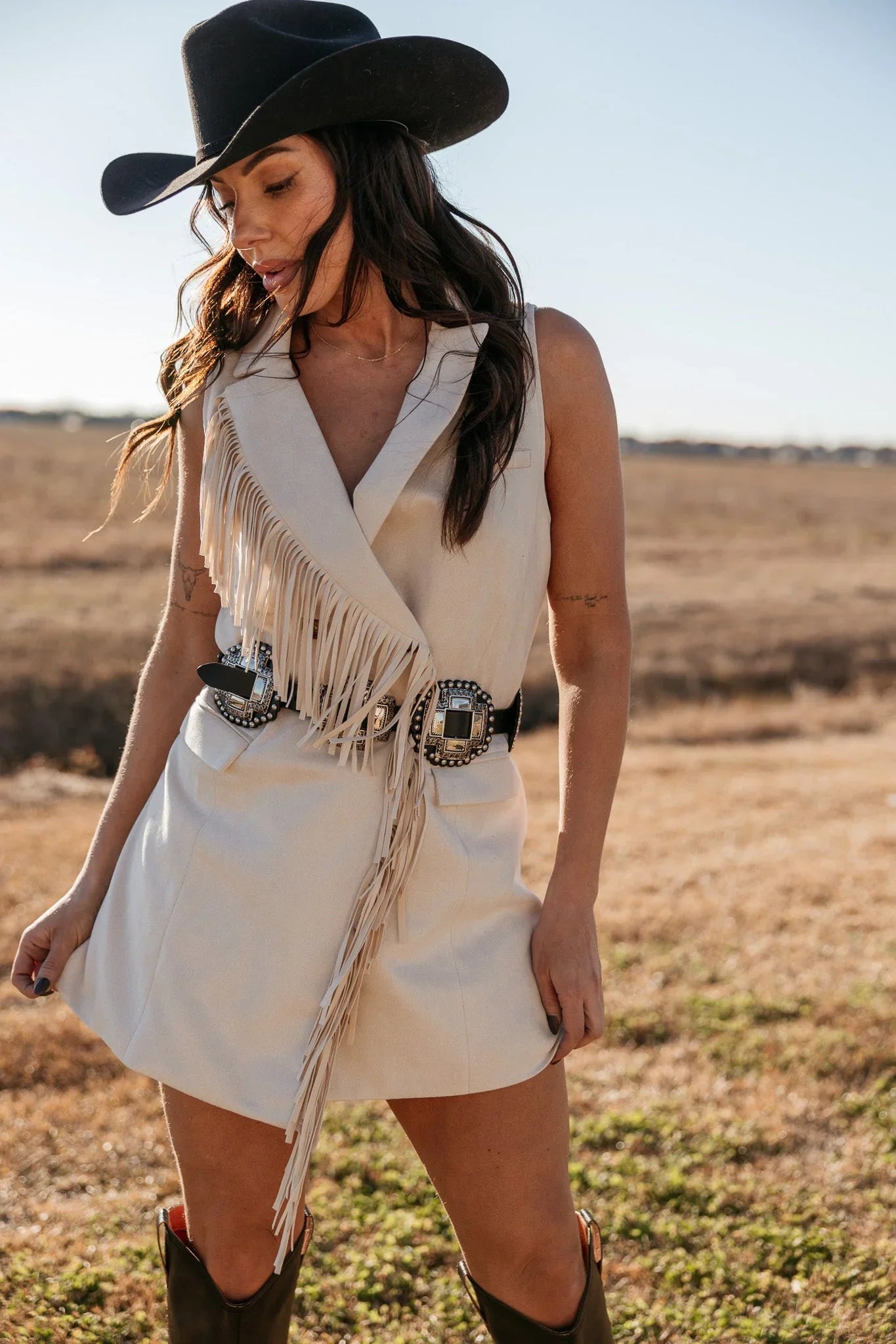 Woman in a beige fringed western vest dress, concho belt, black cowboy hat, and boots outdoors