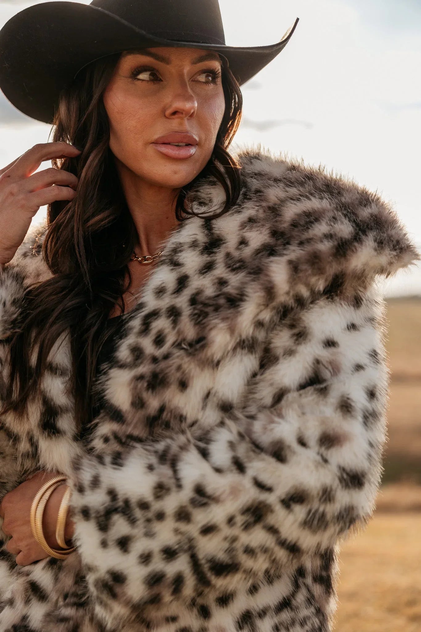 Woman in black cowboy hat and faux fur leopard print coat outdoors, western boho fashion