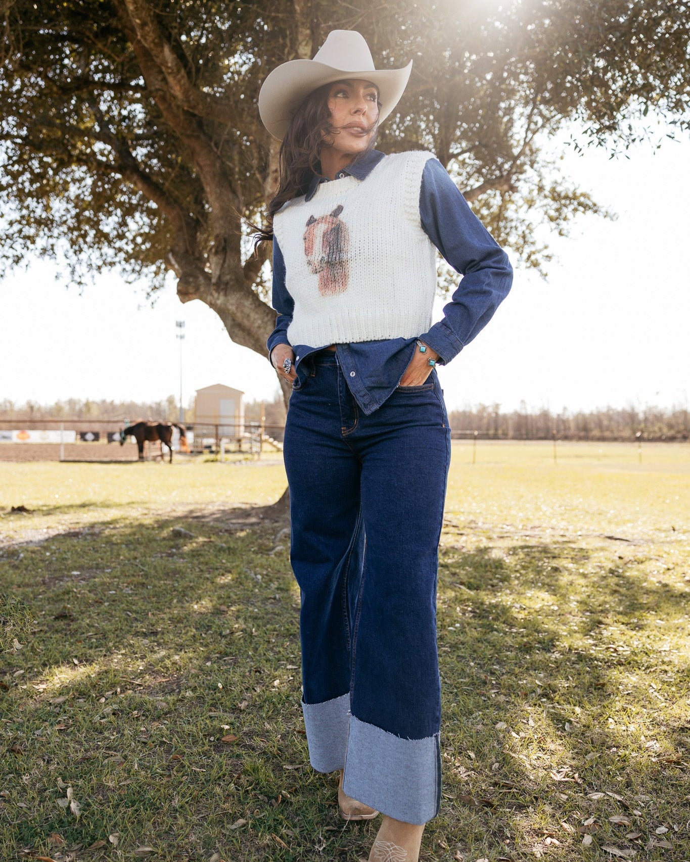 Woman in a cowboy hat, wide-leg jeans, white knit vest and boots, posing outdoors in a western ranch setting