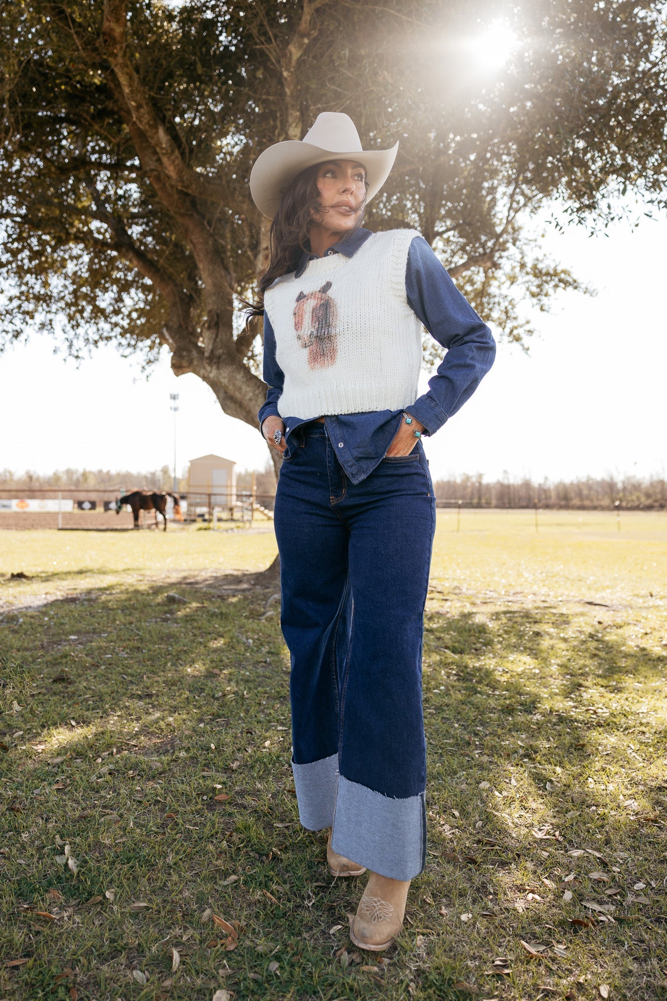 Woman in a cowboy hat, wide-leg jeans, white knit vest and boots, posing outdoors in a western ranch setting