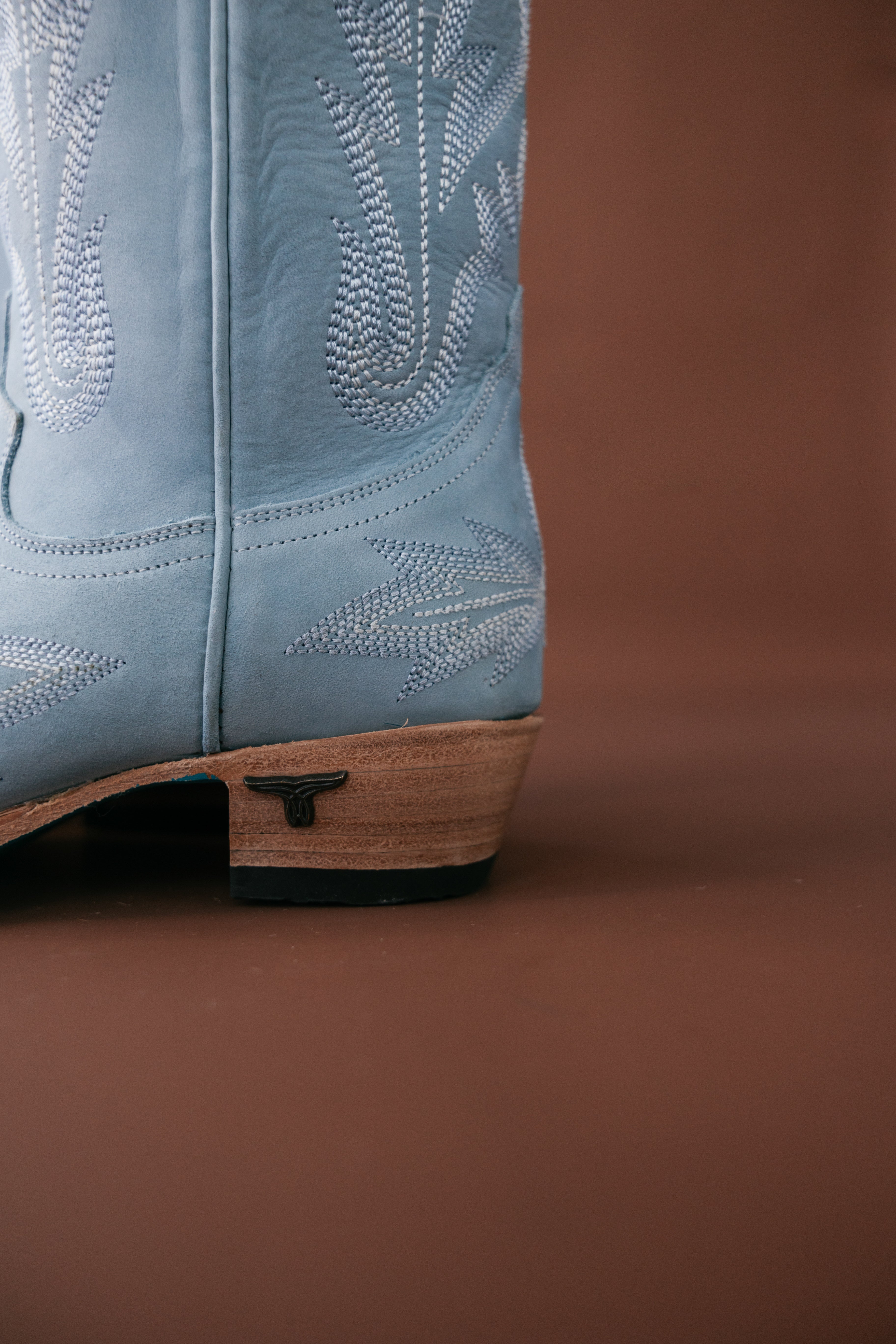 Light blue western cowgirl boot with white stitching and wooden heel on brown background
