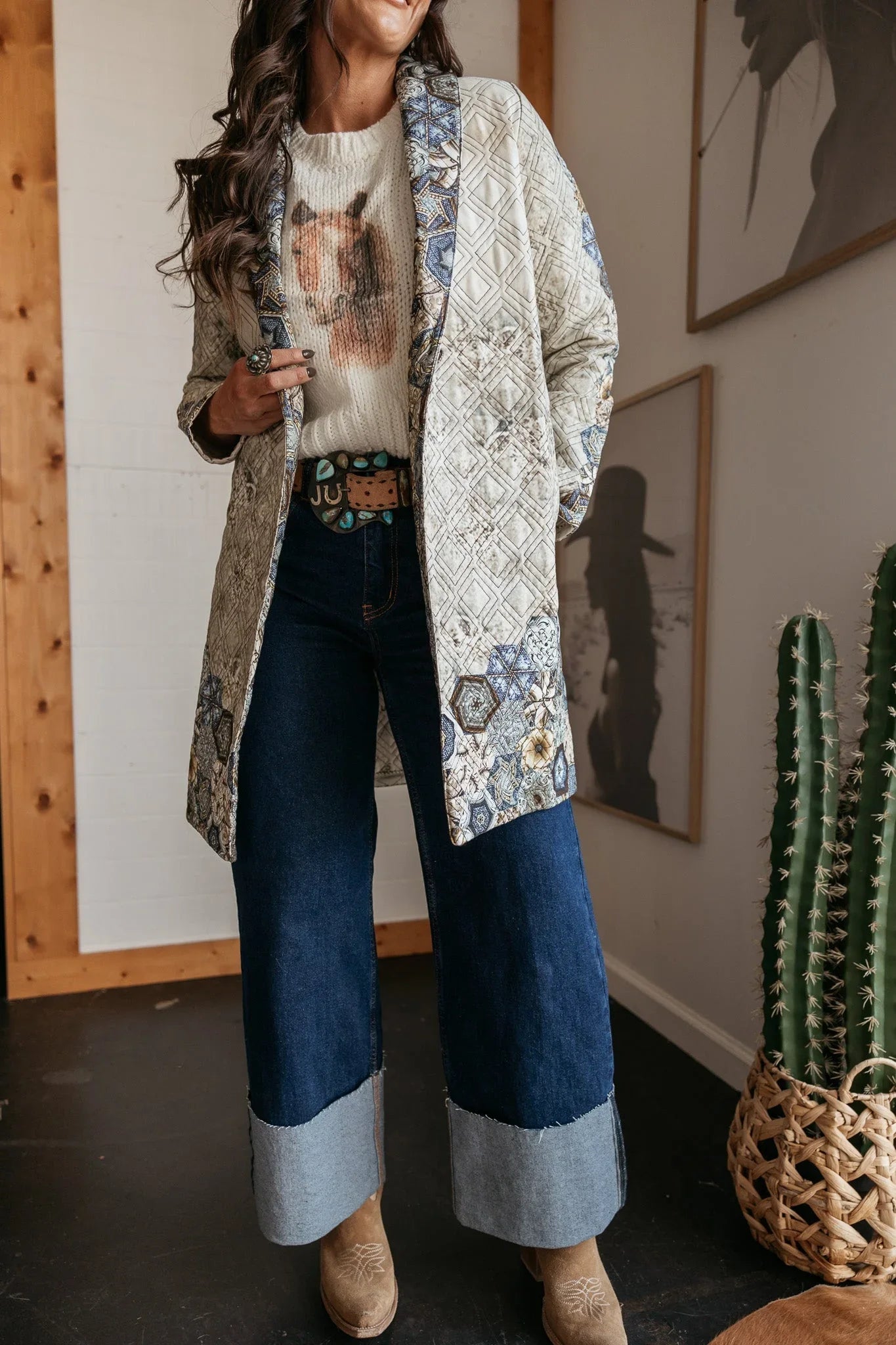Woman in western boho outfit with patterned quilted jacket, horse sweater, wide leg jeans, and cowboy boots