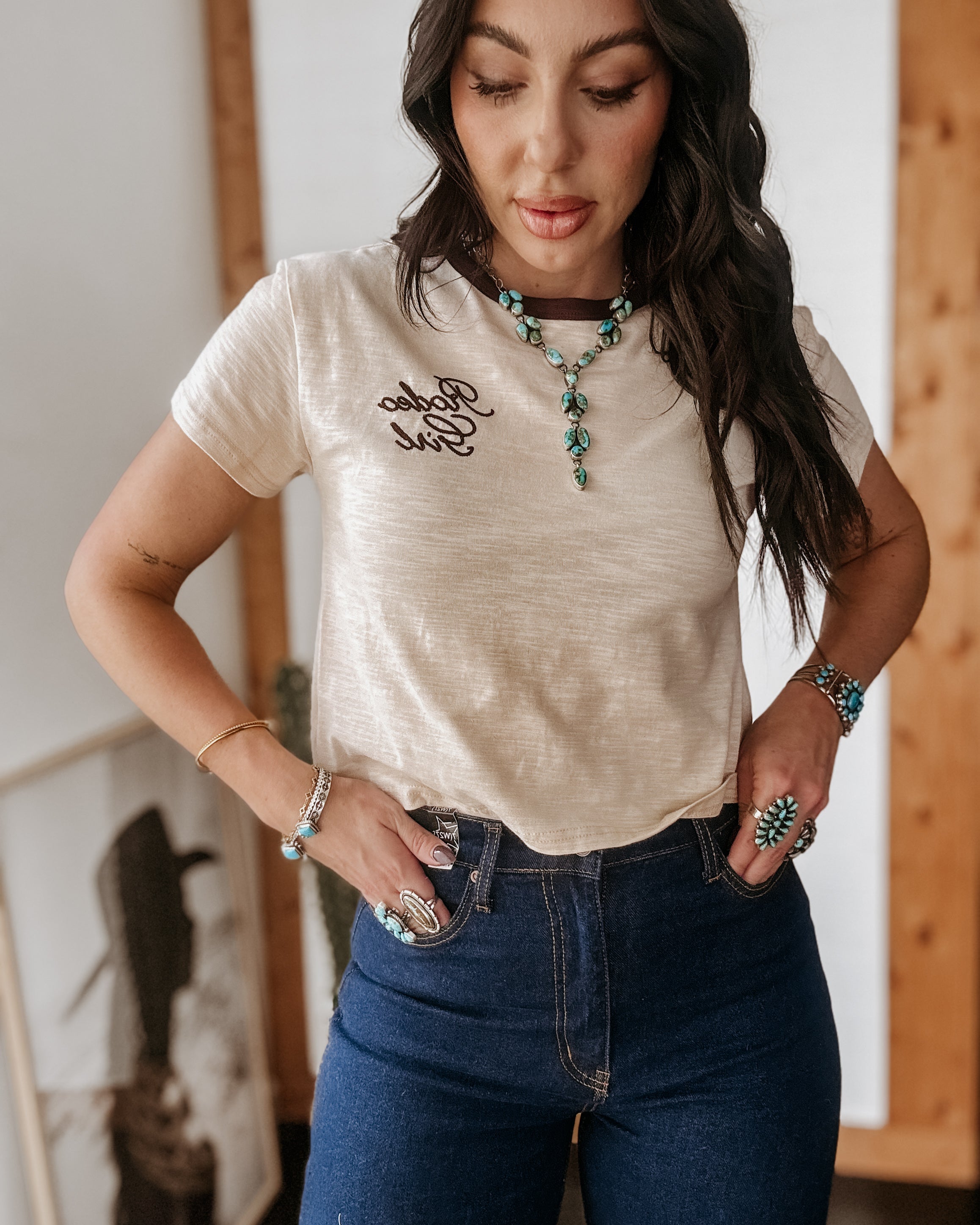 Woman in western boho outfit wearing 'Rodeo Girl' t-shirt, turquoise jewelry, and high-waisted jeans