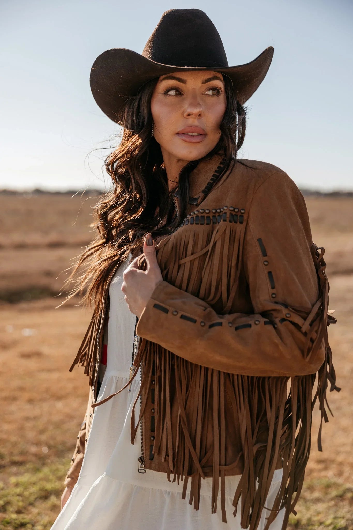 Woman in a brown fringe suede jacket and black cowboy hat in a western outdoor setting