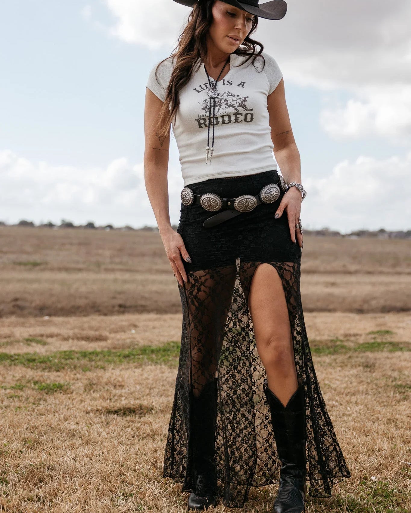 Woman in a cowgirl hat, graphic tee, black lace skirt with slit, western belt, and cowboy boots outdoors