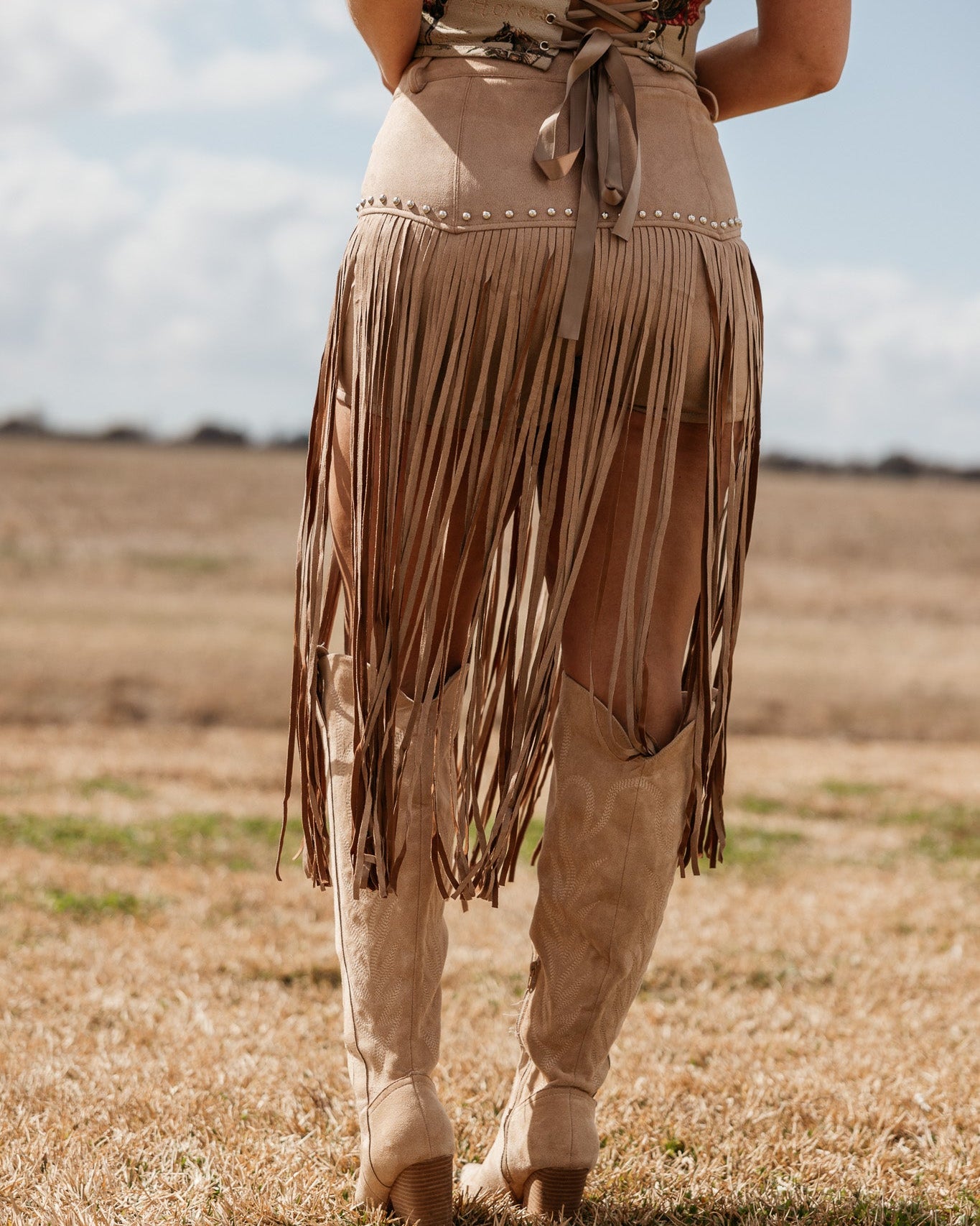 Woman wearing tan fringe western skirt, corset top, and cowboy boots outdoors