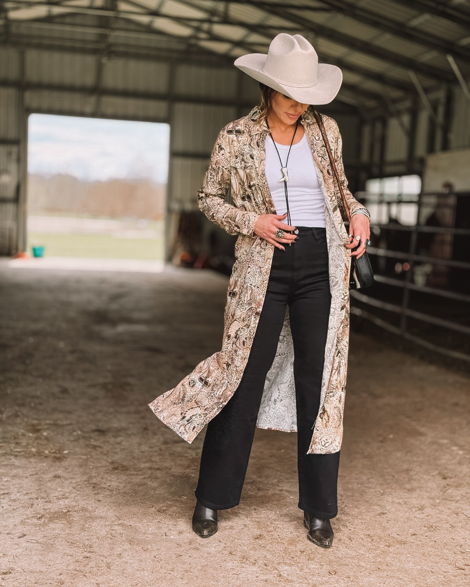 Woman in cowgirl hat, paisley duster, white top, and black pants in barn, western fashion