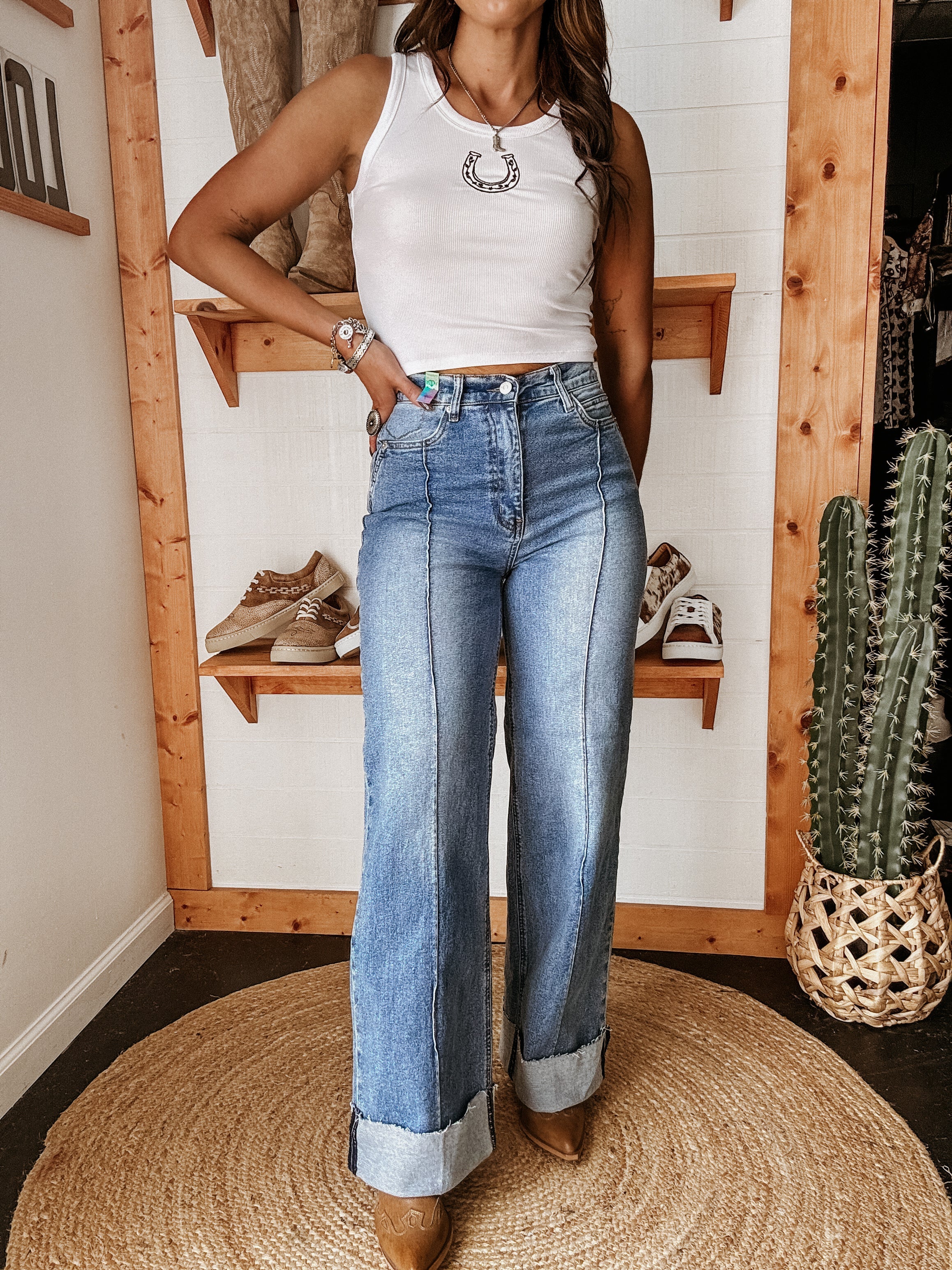 Woman in western boho outfit with white tank top, horseshoe necklace, high-waisted wide leg jeans, and cowgirl boots standing indoors