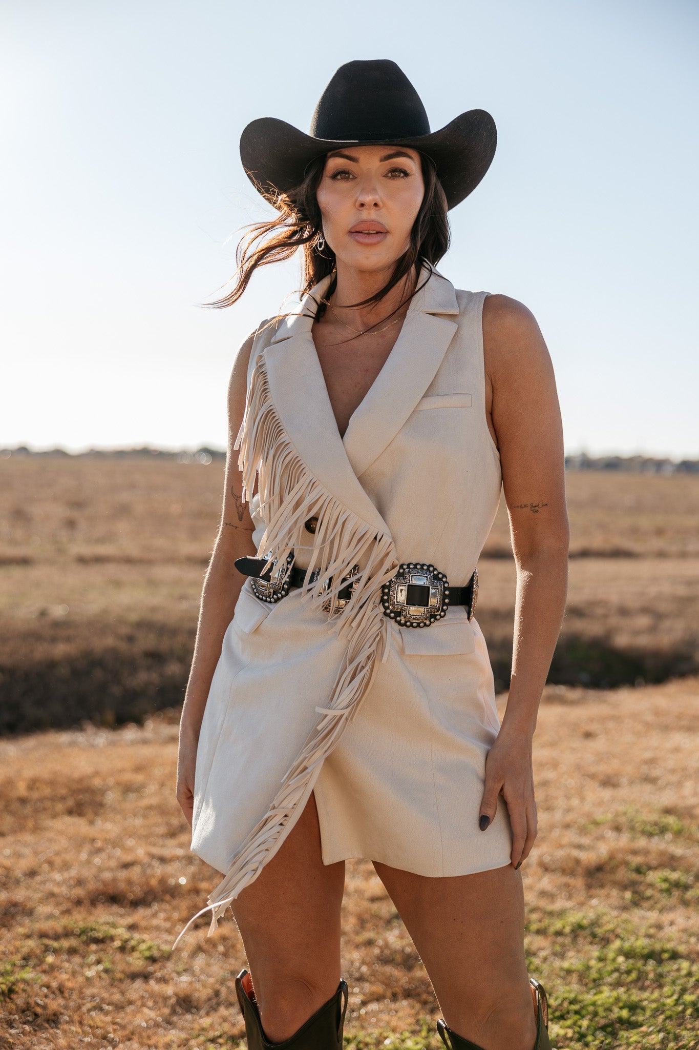 Woman in sleeveless beige fringe western dress, black cowboy hat, western belt, outdoor setting
