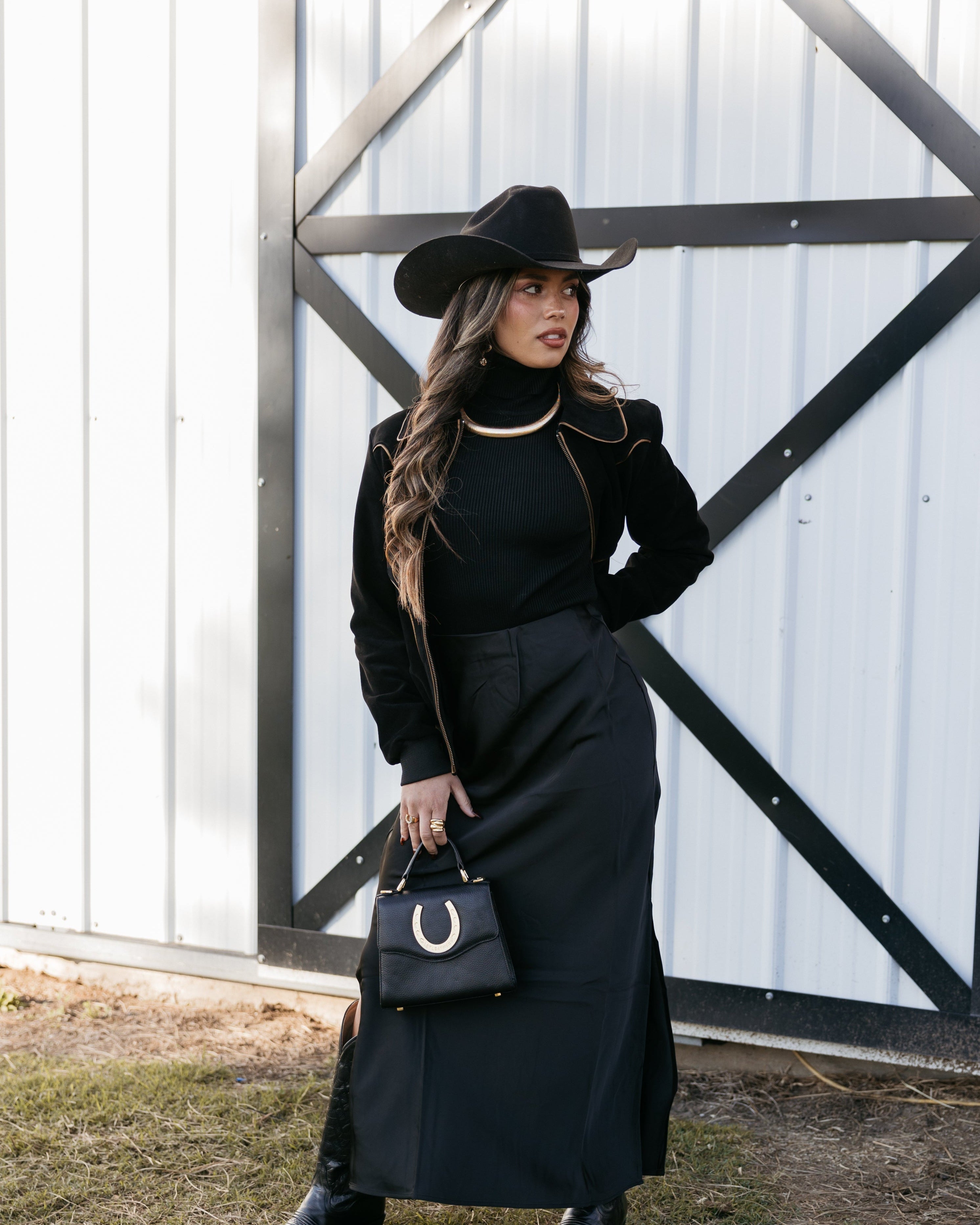 Woman in black cowgirl outfit with wide-brim hat, maxi skirt, and horseshoe purse standing by barn